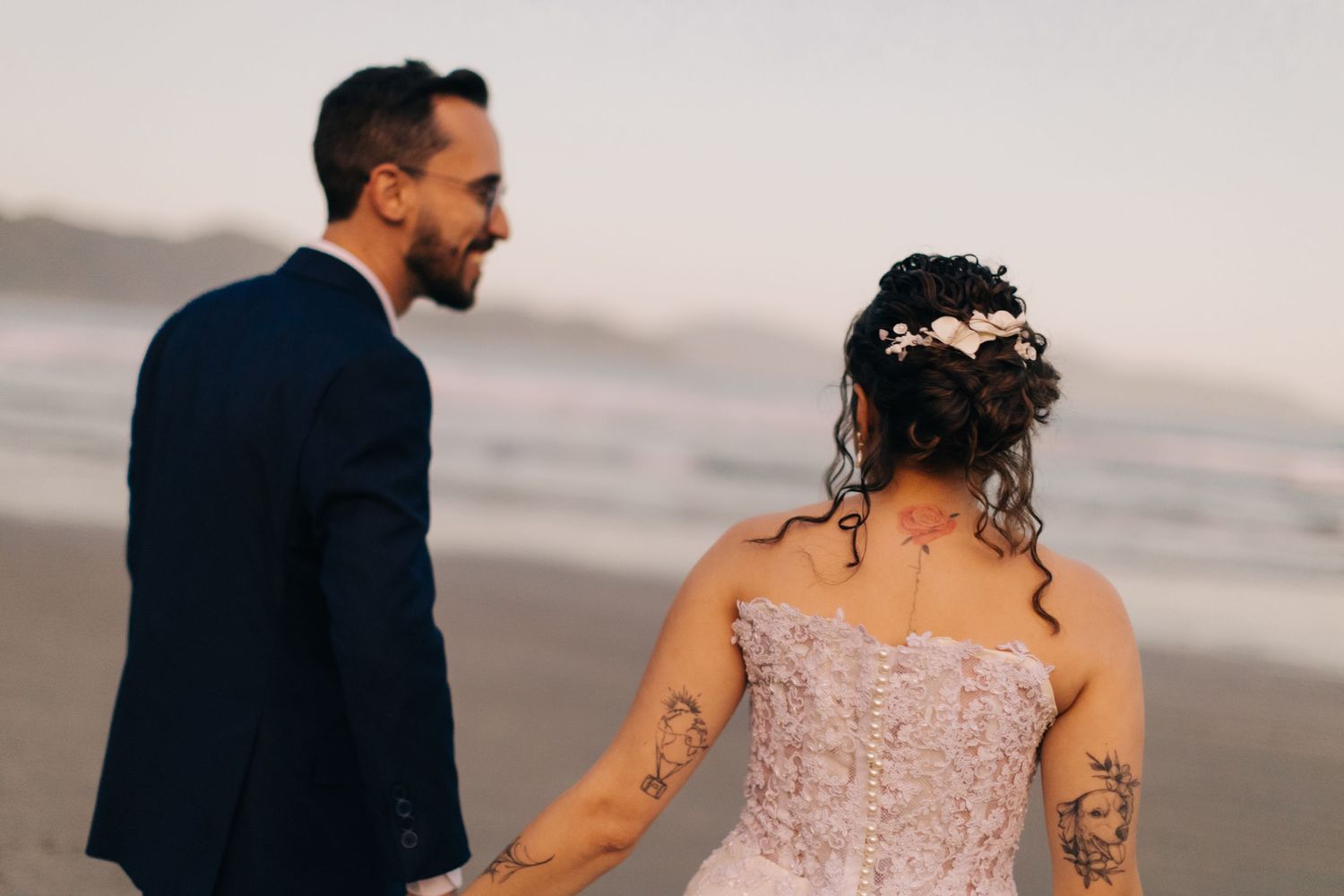 A couple in wedding attire walking along a misty beach at sunset.