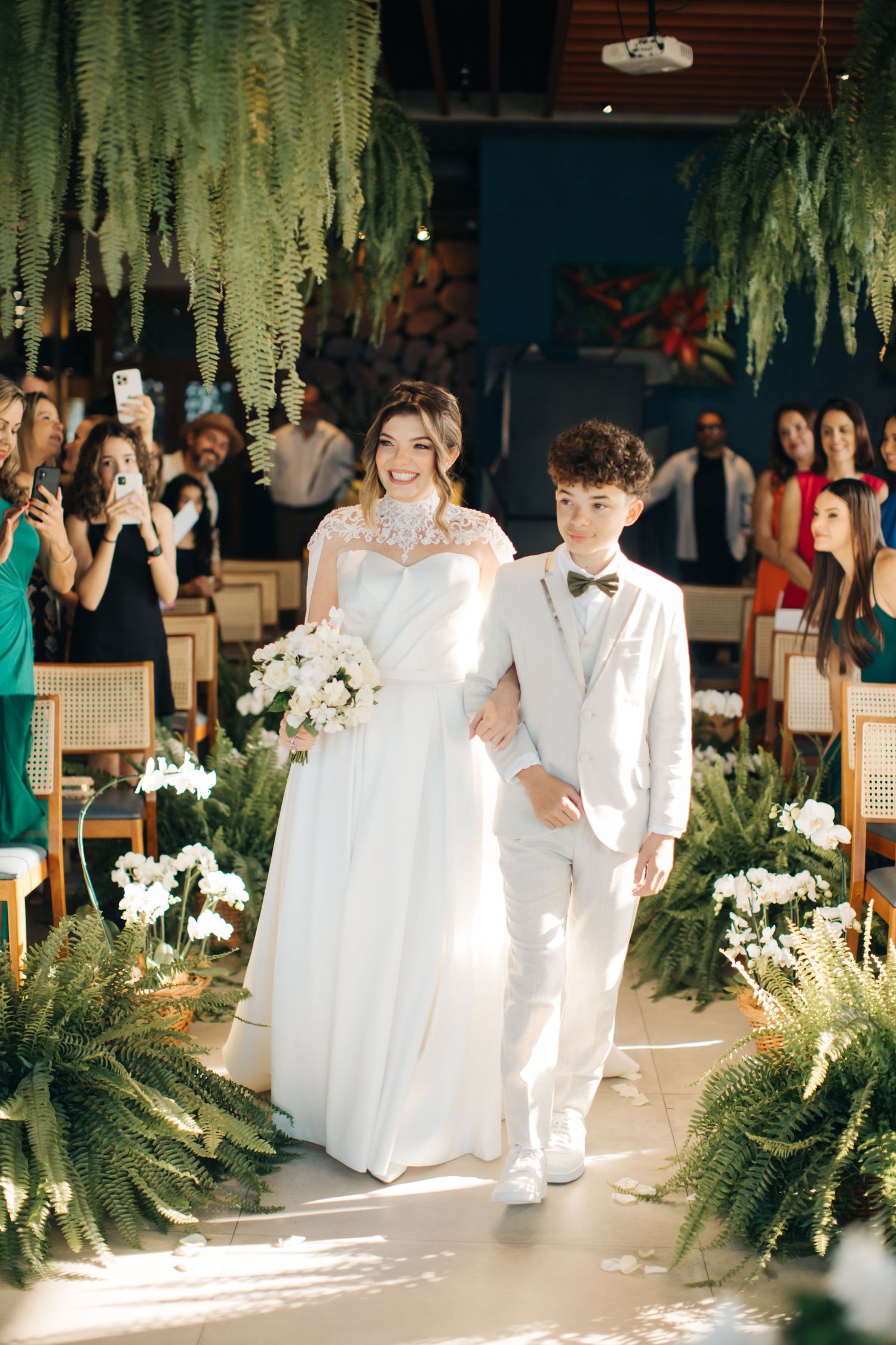 Wedding ceremony setup with white-clad attendants walking down an aisle decorated with ferns.