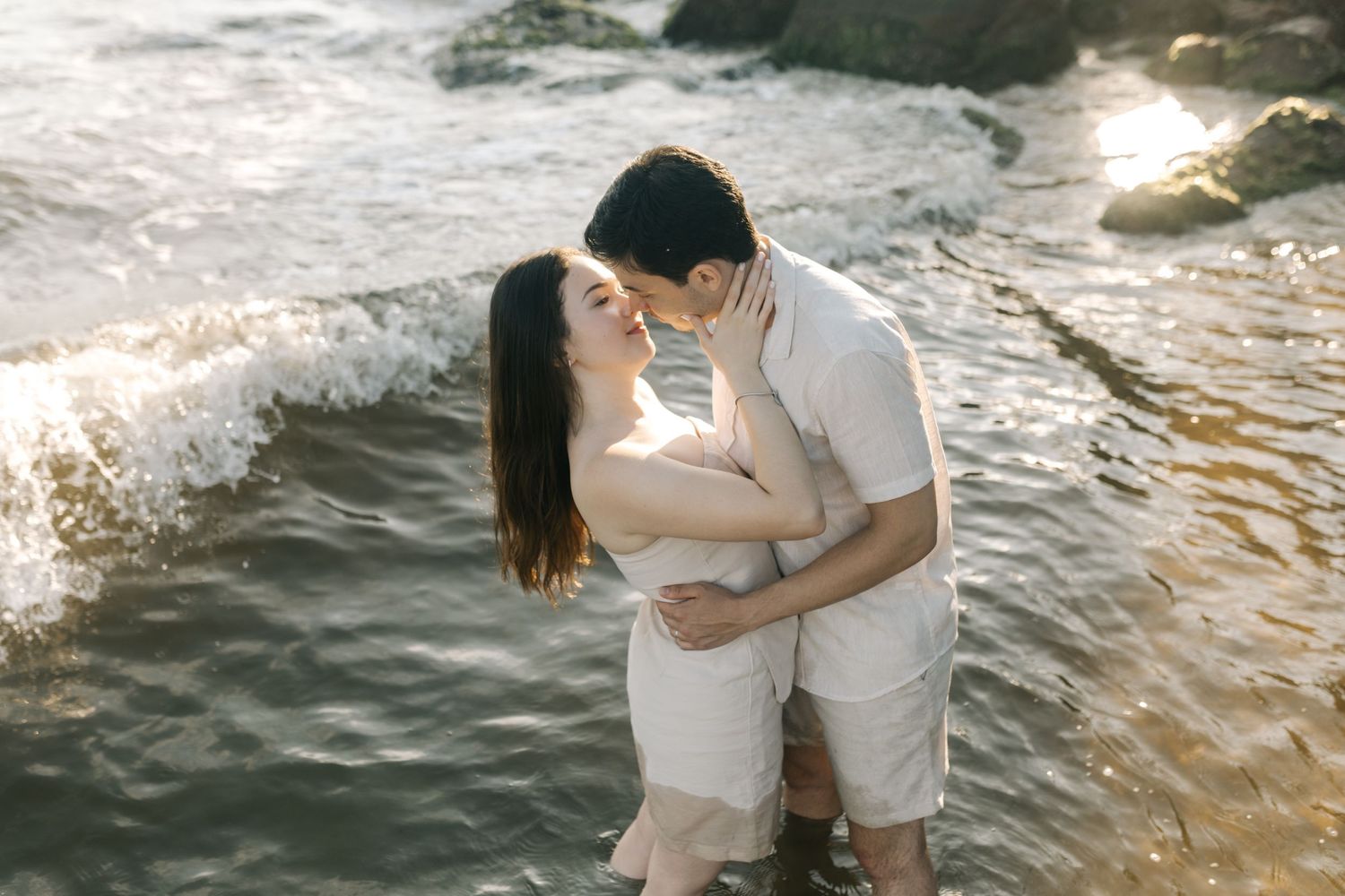 A couple stands in shallow water at sunset sharing intimate moments on a rocky beach.
