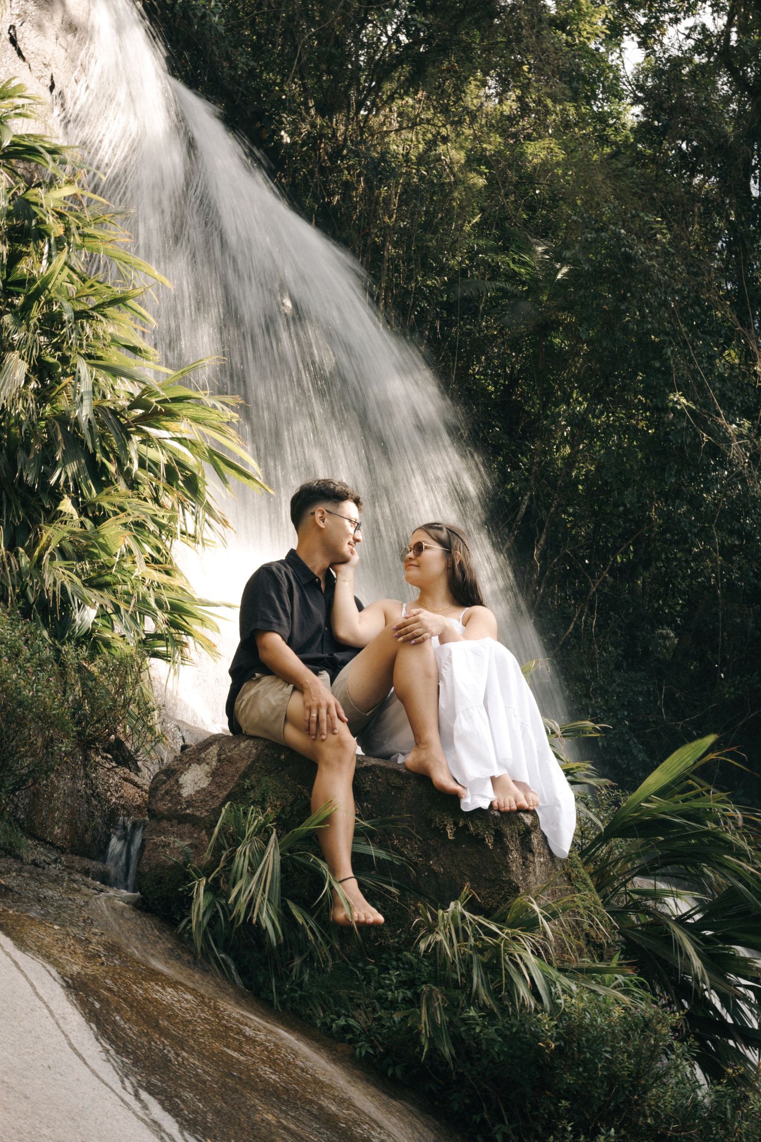 Couple sharing a peaceful moment on a rock ledge with a dramatic waterfall backdrop.