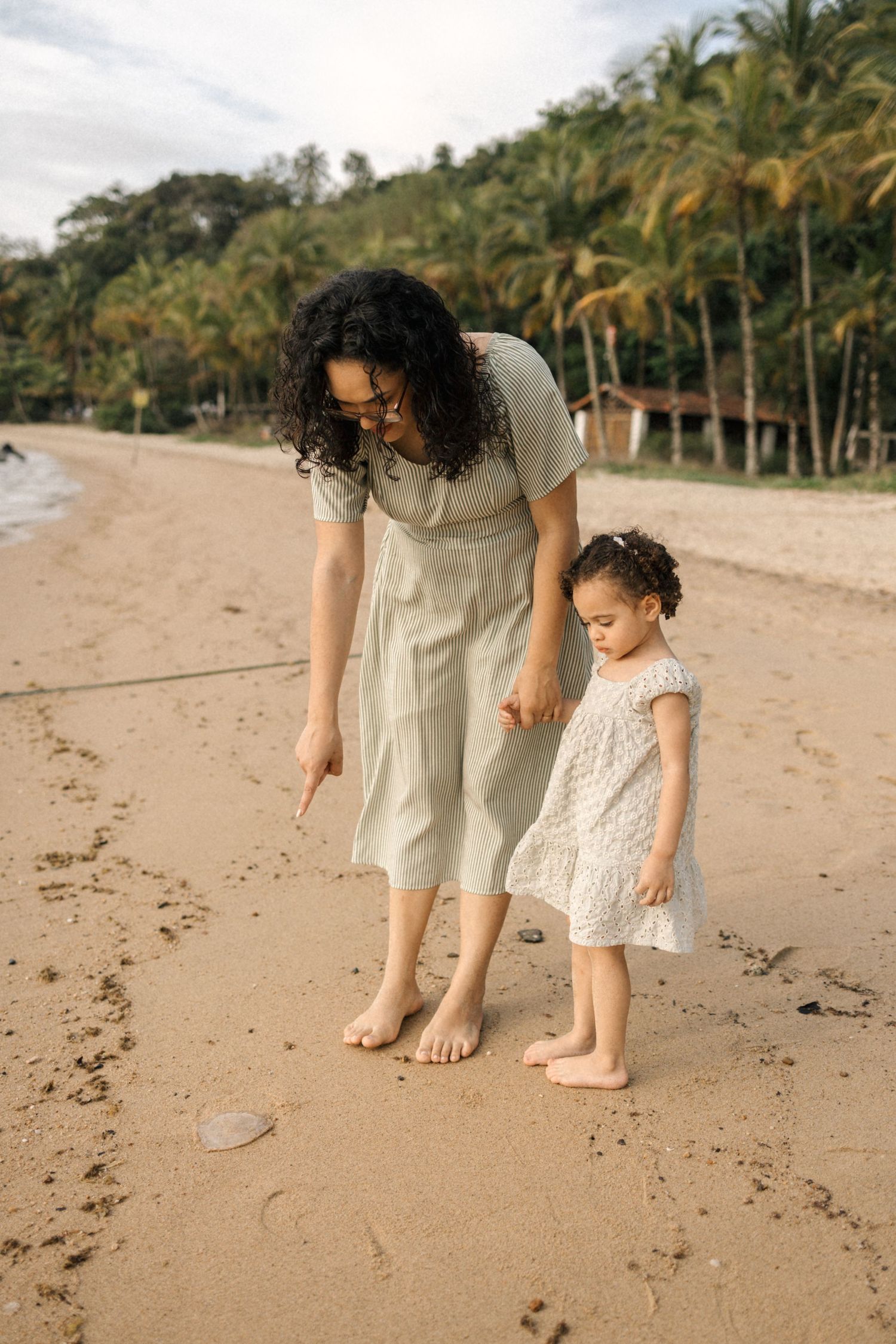 Two people explore a tropical beach with palm trees while barefoot in light summer dresses.