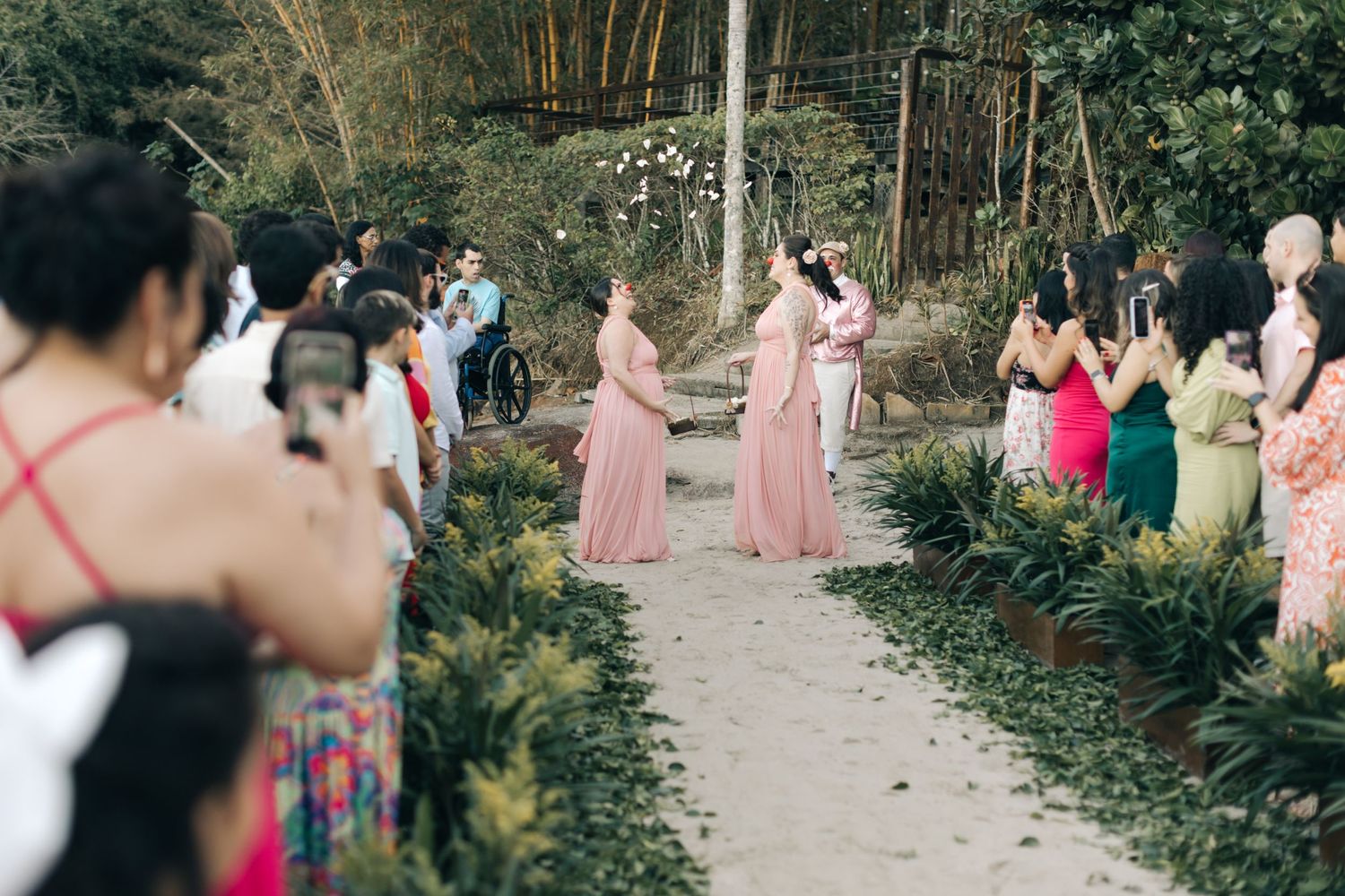 Bridesmaids in pink dresses pose while guests take photos in a tropical garden setting.