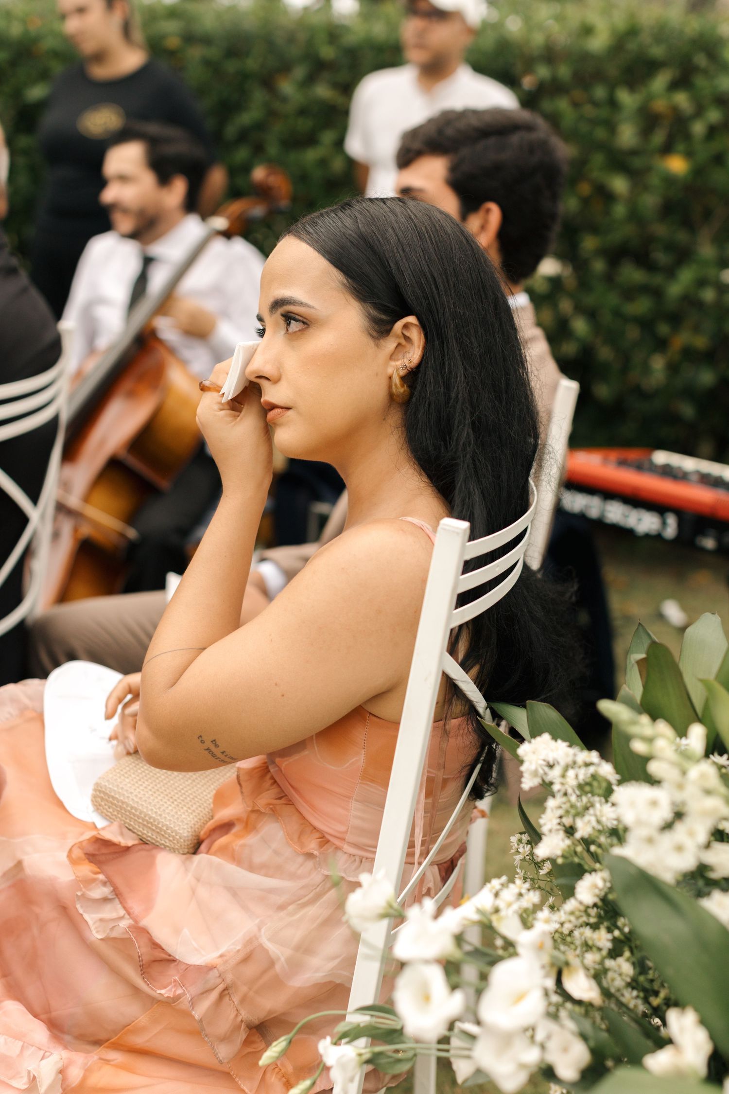 Wedding guest in a pink dress sitting near musicians with cello at an outdoor event.
