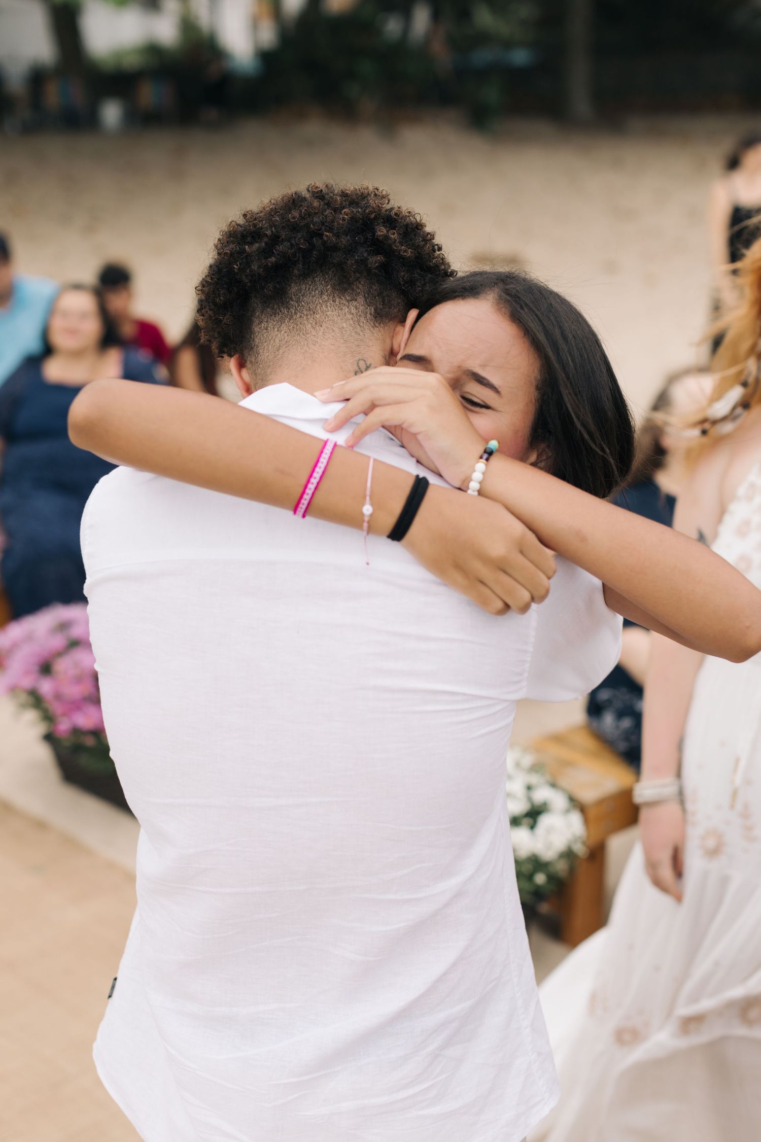 Two people sharing an emotional embrace while wearing white clothing during a beach ceremony.