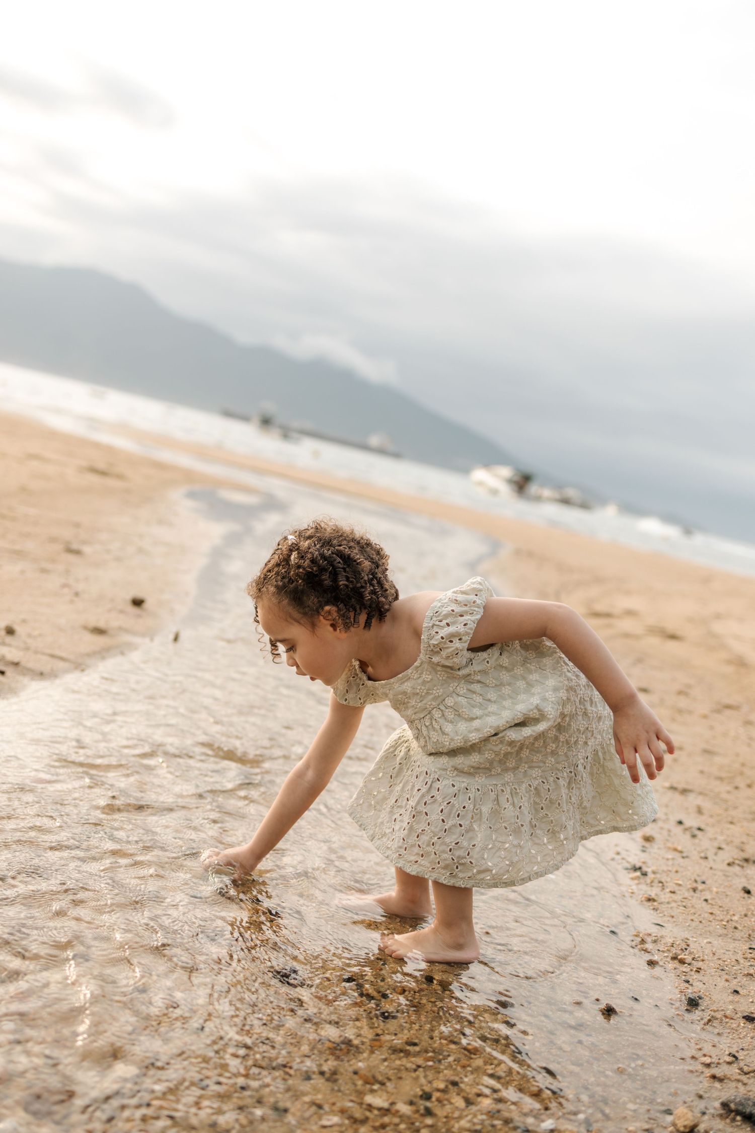 A small child in a floral dress bends down to play in the shallow water at the beach.