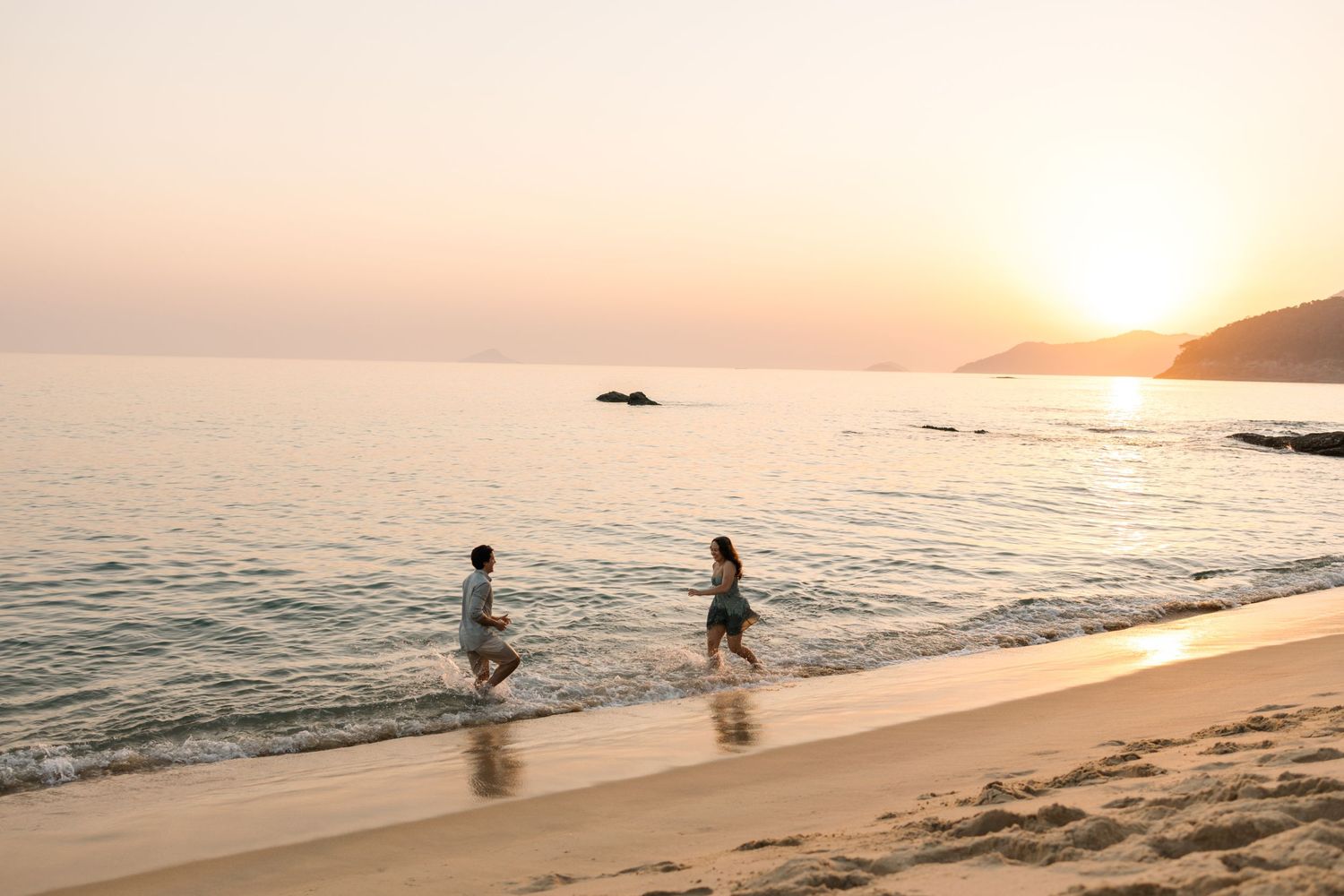 People playing in gentle ocean waves at sunset along a peaceful sandy beach.