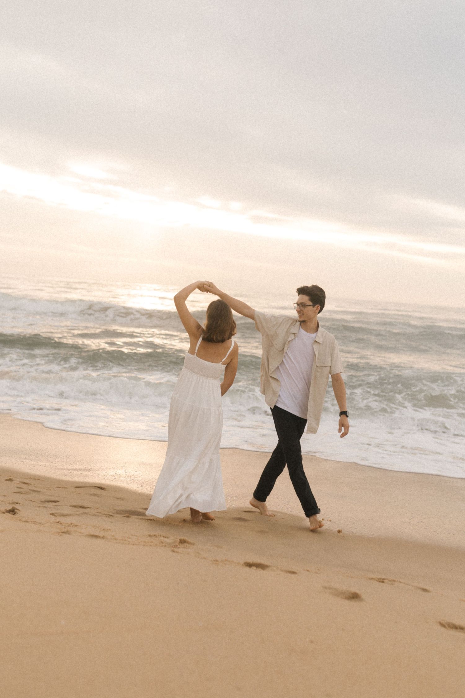 A couple dances playfully on the beach during a golden sunset with waves in the background.