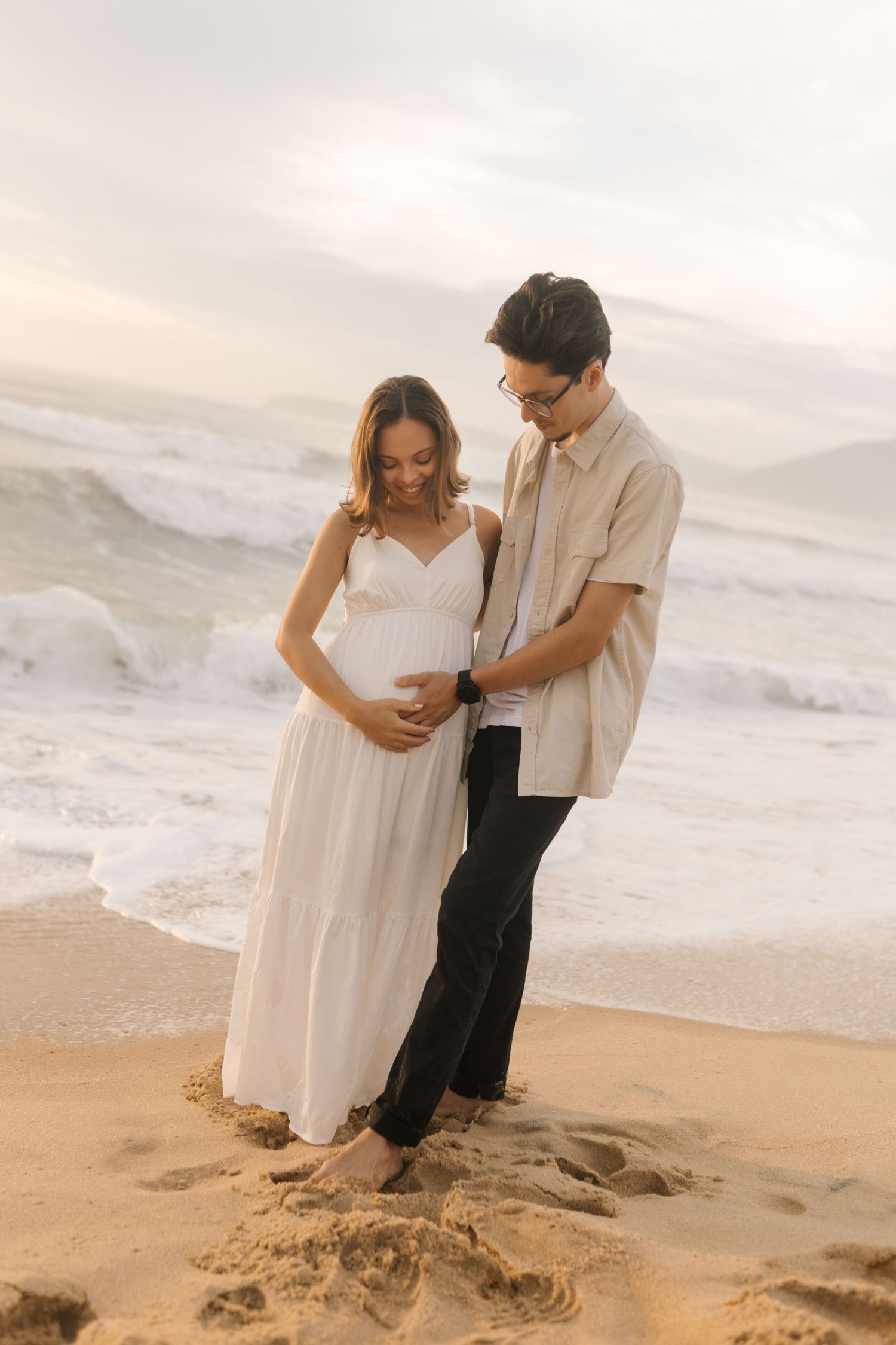 A couple in light clothing shares a tender moment on a rocky beach with ocean waves in the background.