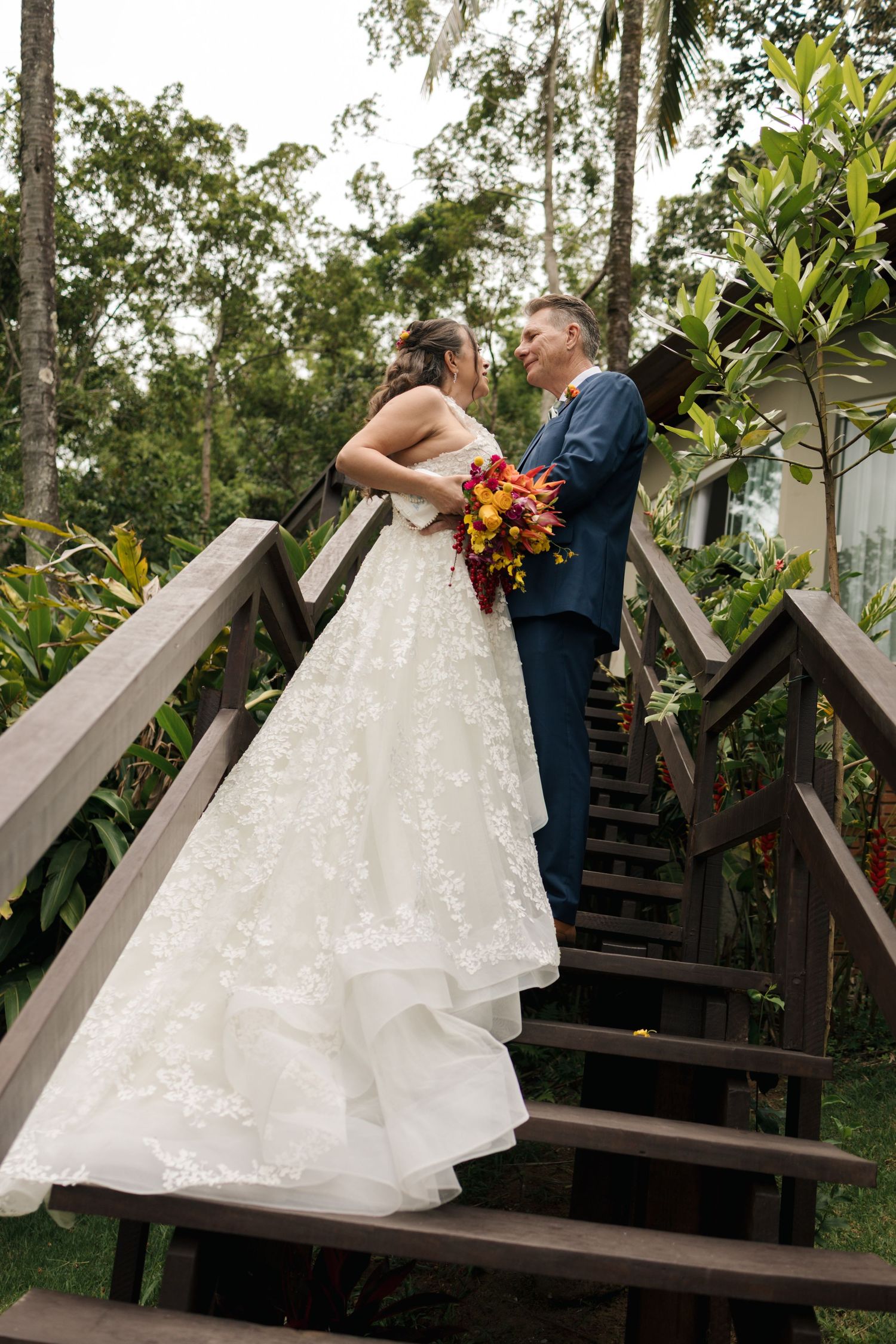 A couple poses on wooden stairs surrounded by lush tropical foliage during their wedding.