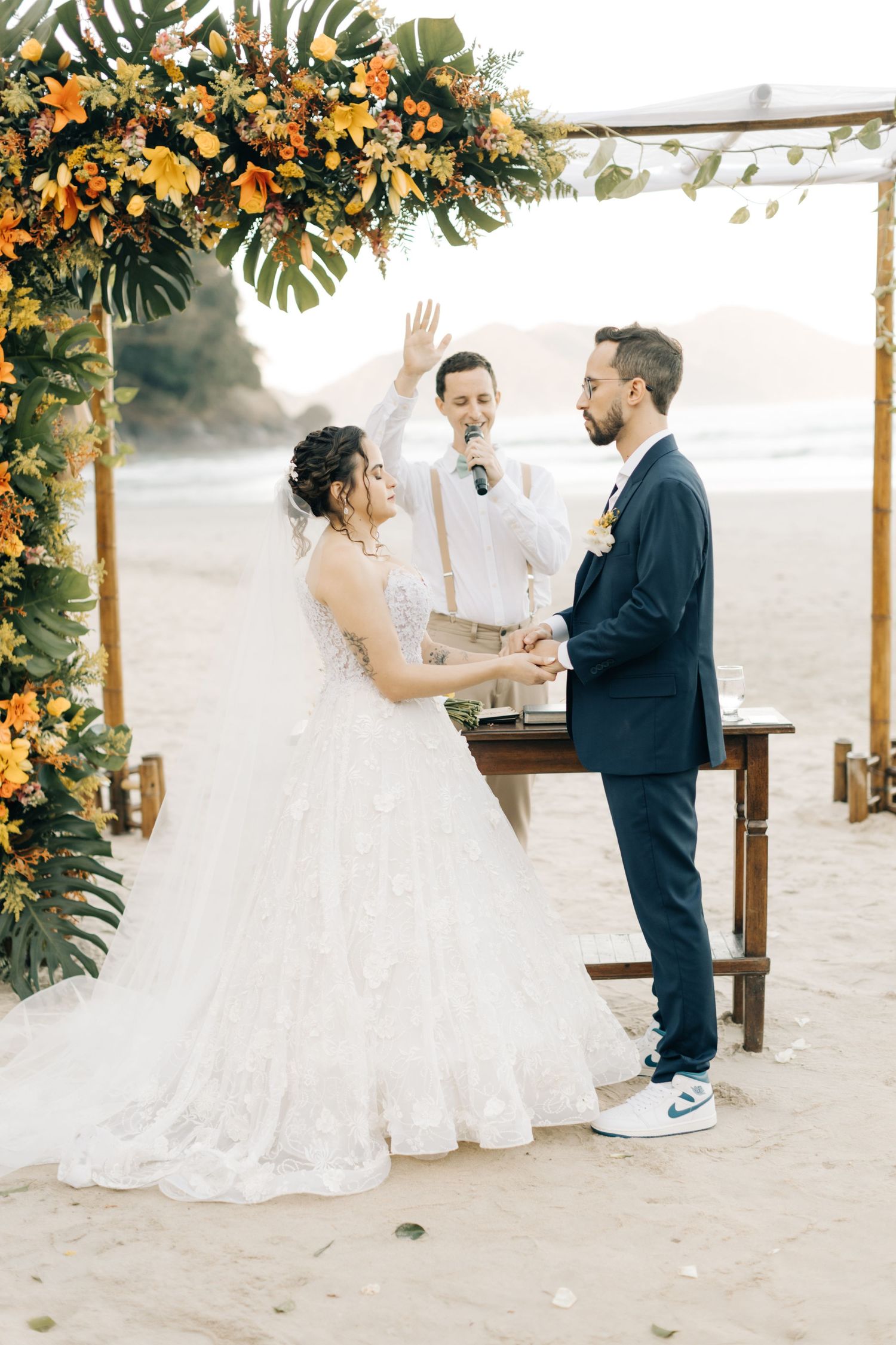 Beach wedding ceremony under floral arch with orange blooms and tropical leaves at golden hour.