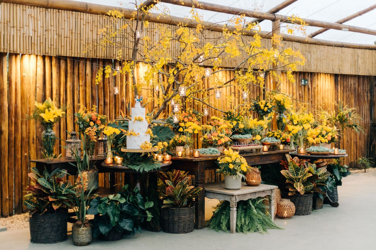Rustic wedding dessert table with bamboo backdrop decorated with yellow florals and hanging lights.