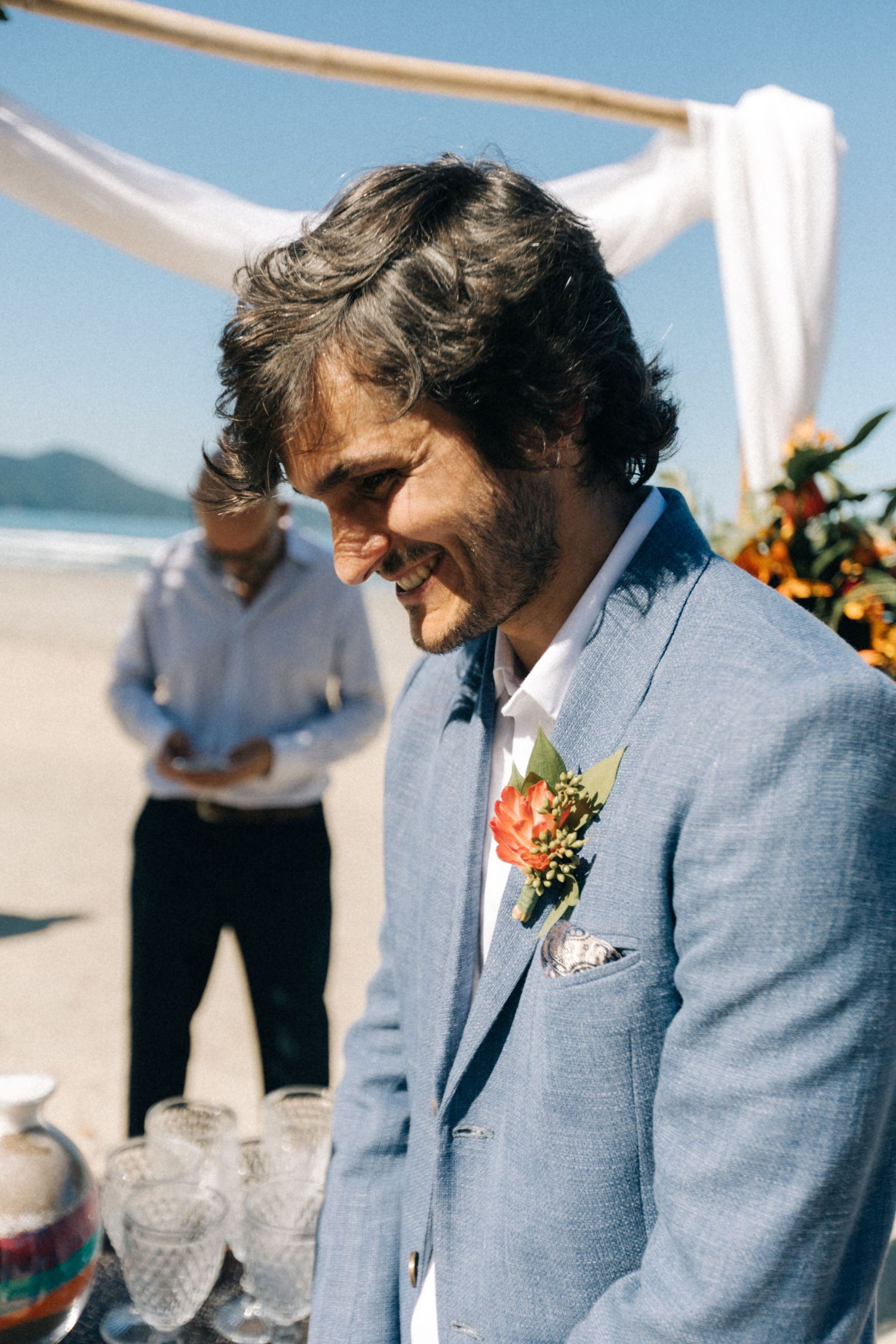 A person in a light blue suit stands on a sunny beach under white fabric draped archways.