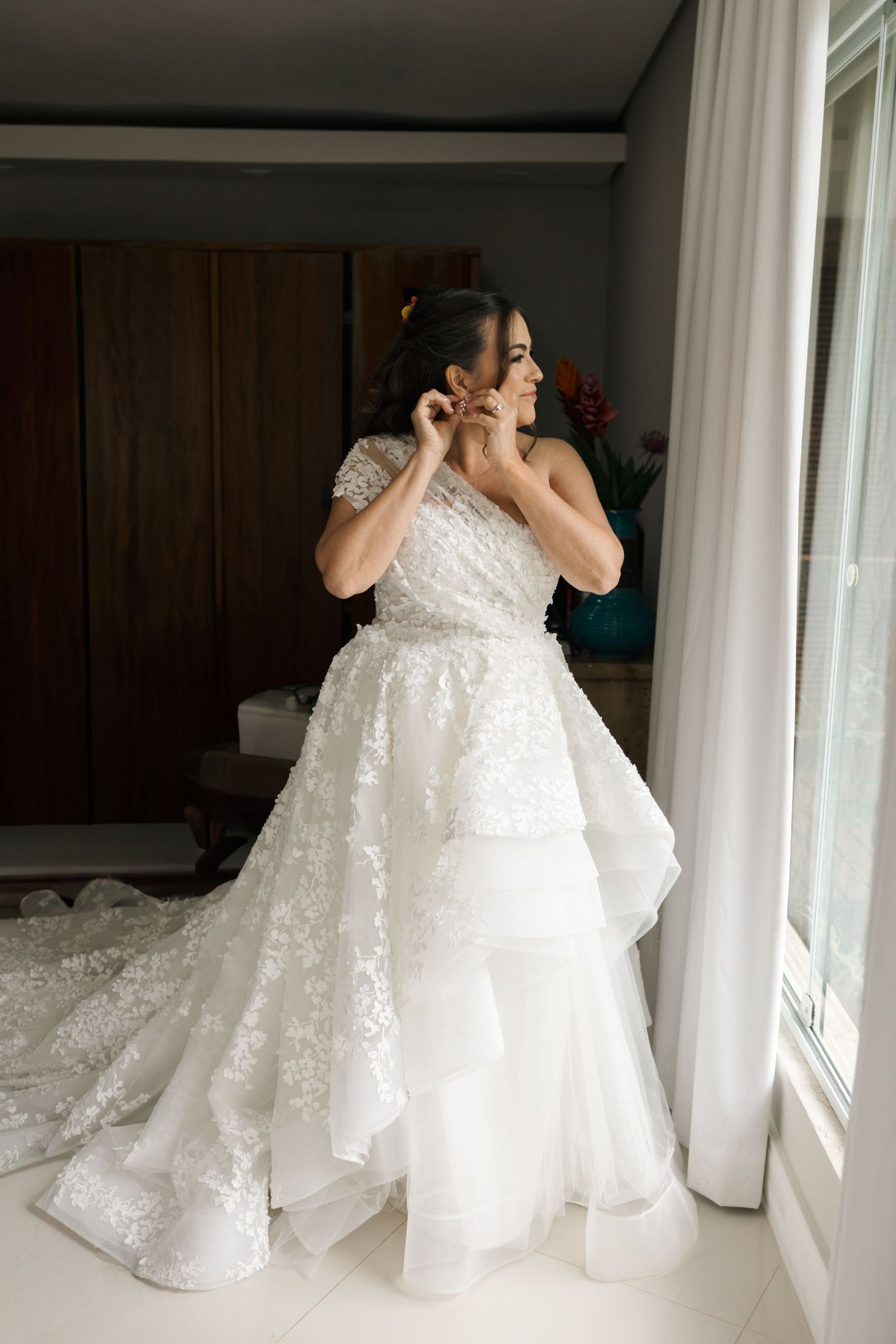 An elegant white wedding gown with intricate lace details and full skirt being fitted in a dressing room.