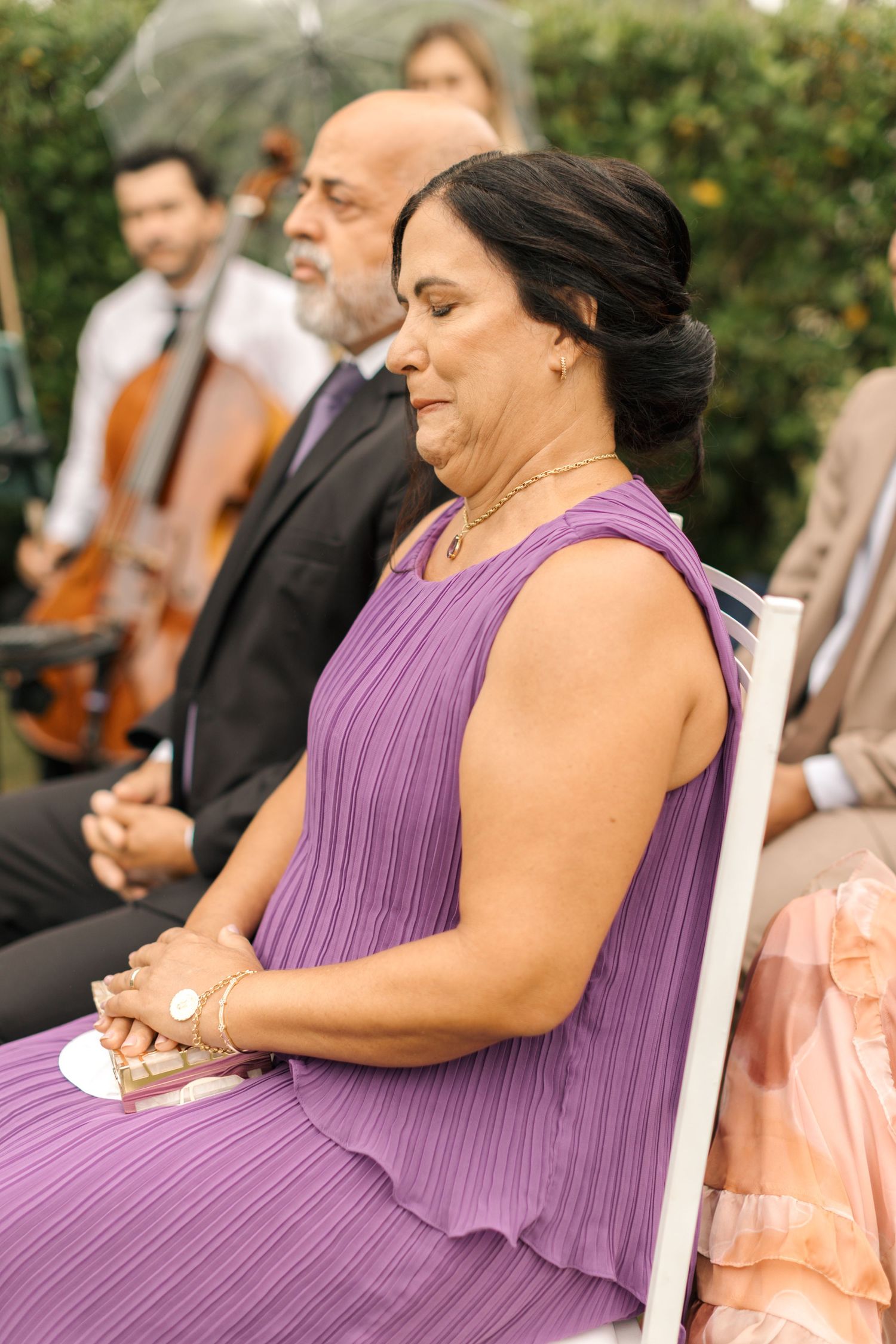 A woman in a purple pleated dress sits attentively during an outdoor event.