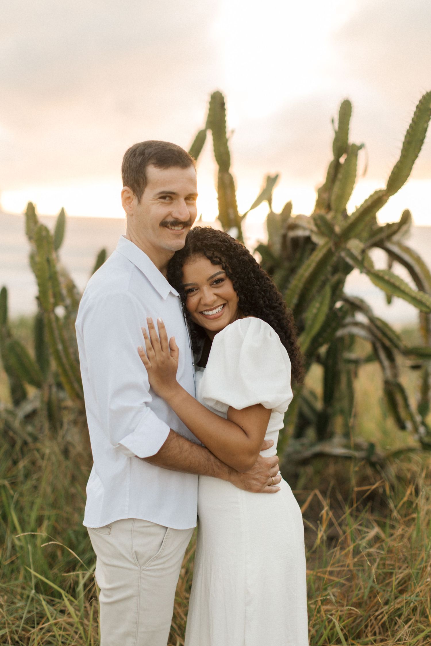 A couple poses together outdoors in white outfits with tall cactus plants in the background during golden hour.