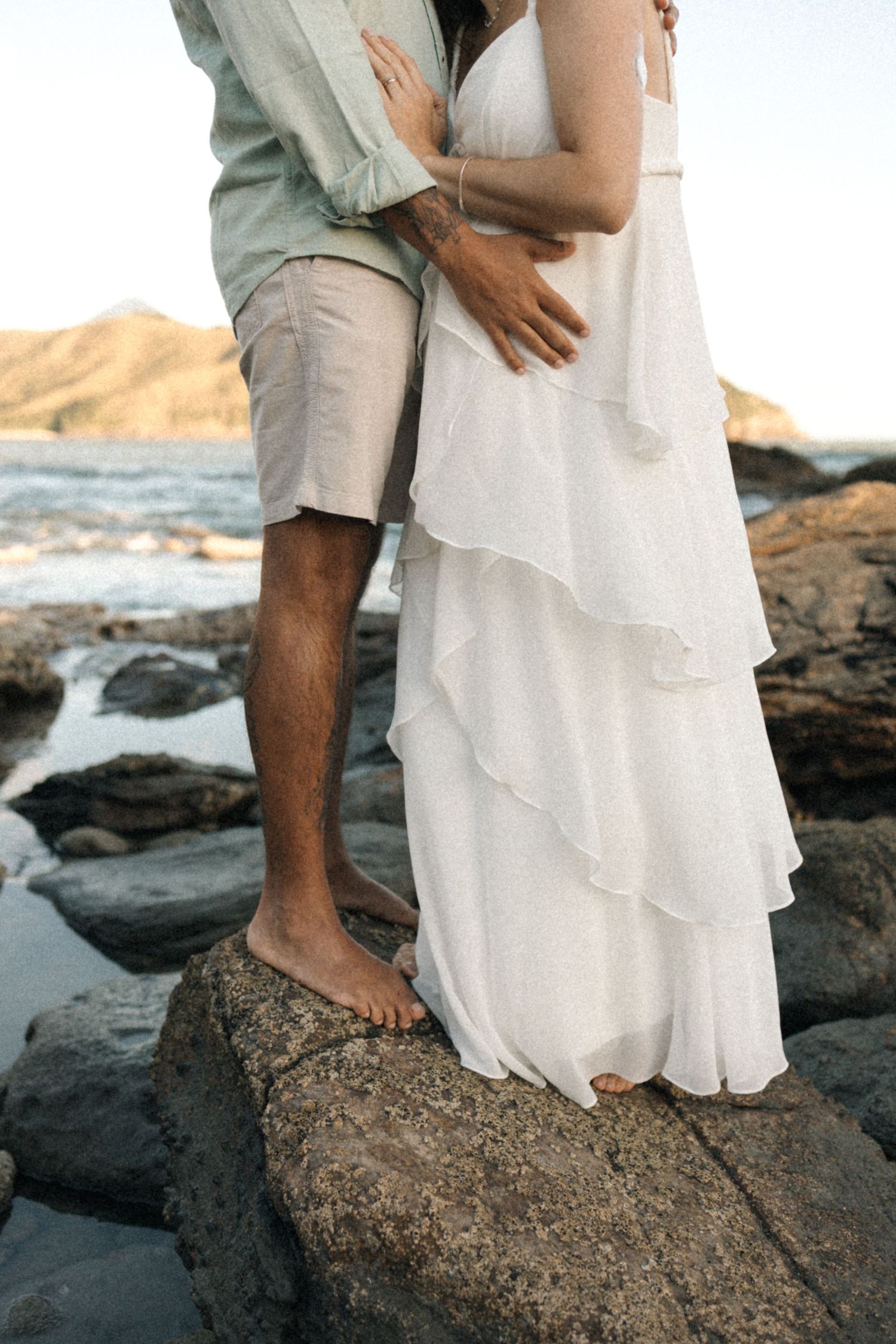 Two people stand close together on rocky coastal terrain wearing white and light colored clothing.