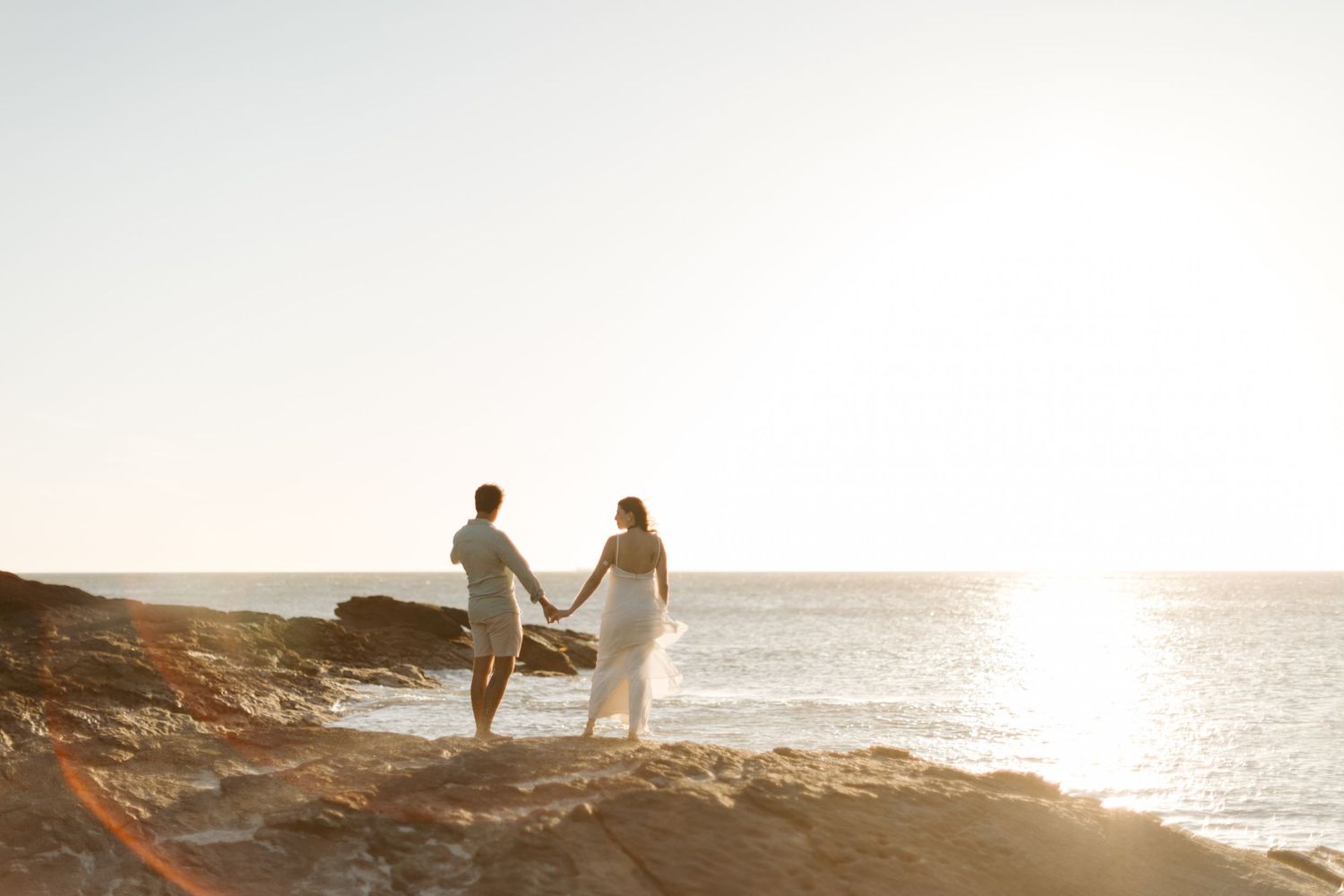Silhouetted couple walking hand in hand along a rocky coastline at sunset.
