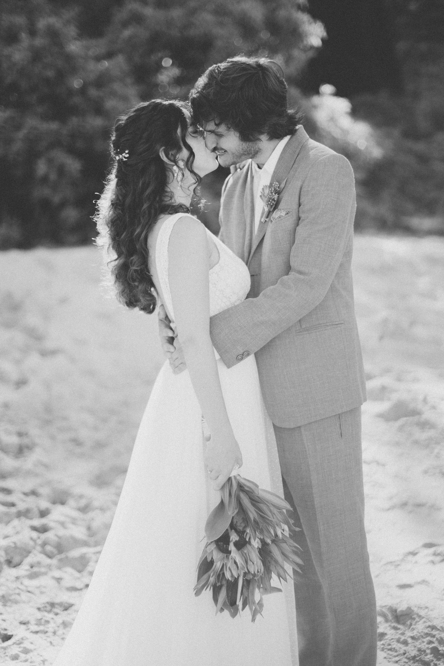 Black and white photo of couple embracing on beach with tropical flower bouquet.
