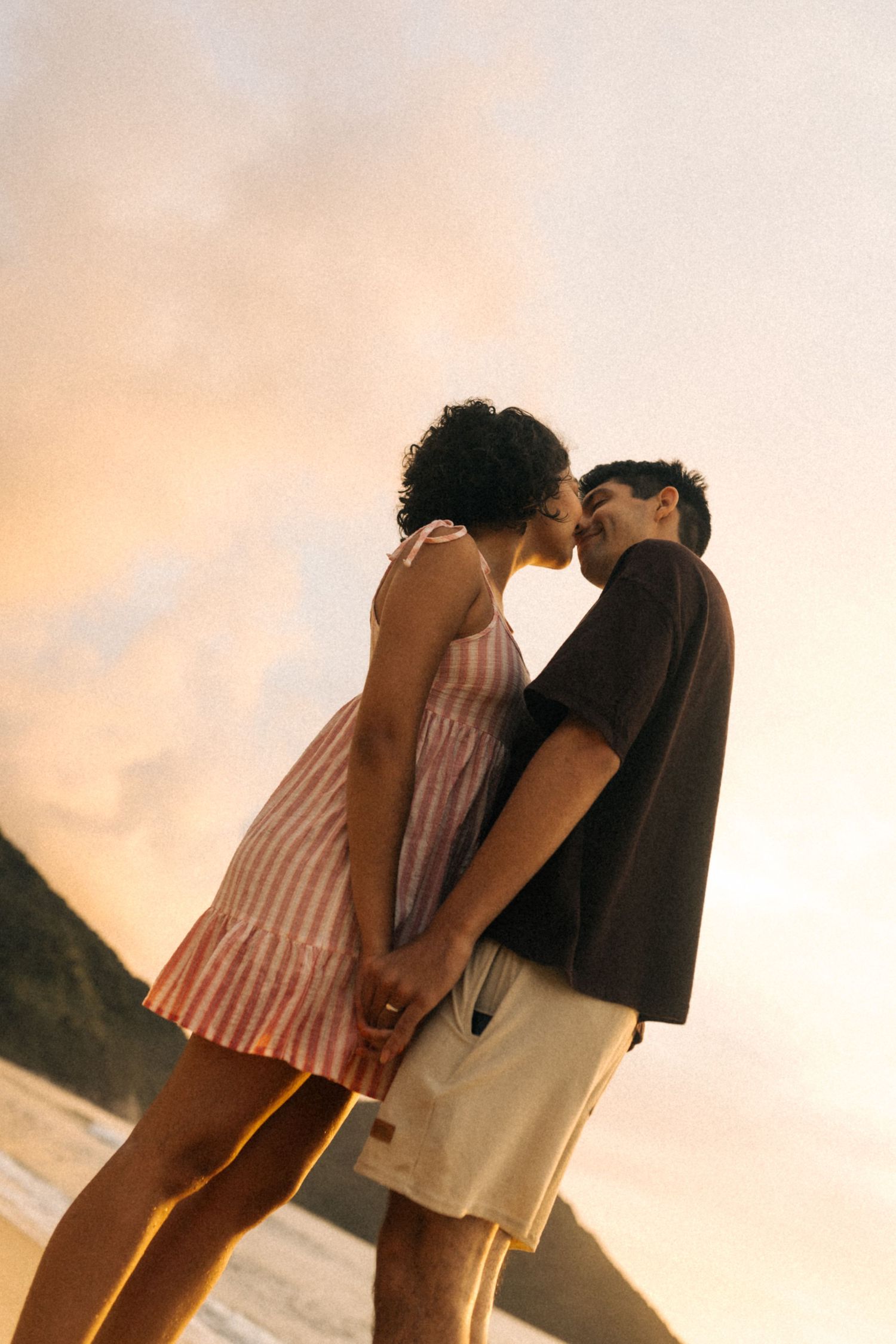 Um casal em trajes de verão compartilha um momento íntimo ao pôr do sol em uma praia, recortado contra um céu dourado.