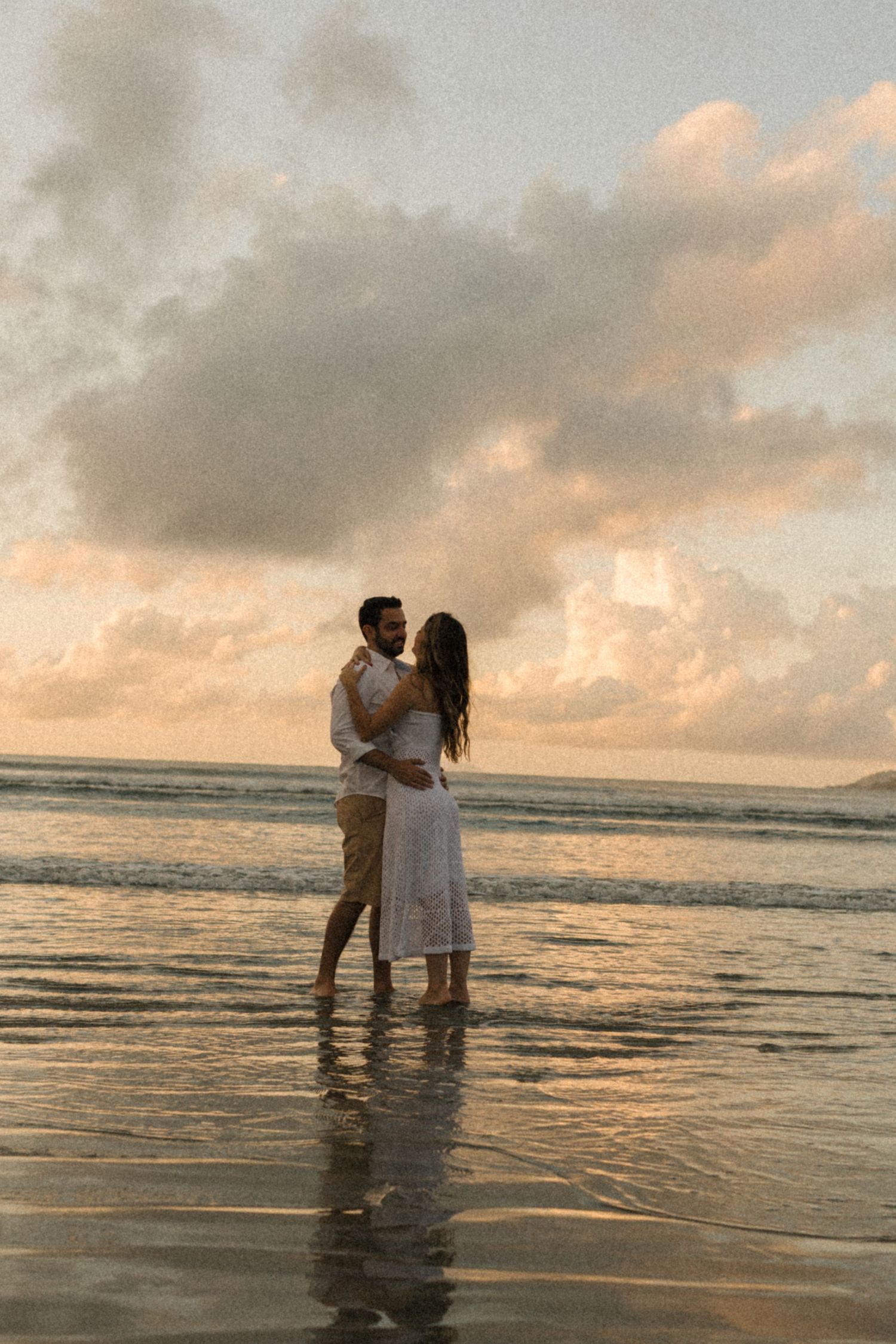 A couple stands together in shallow ocean water during a peaceful sunset moment.