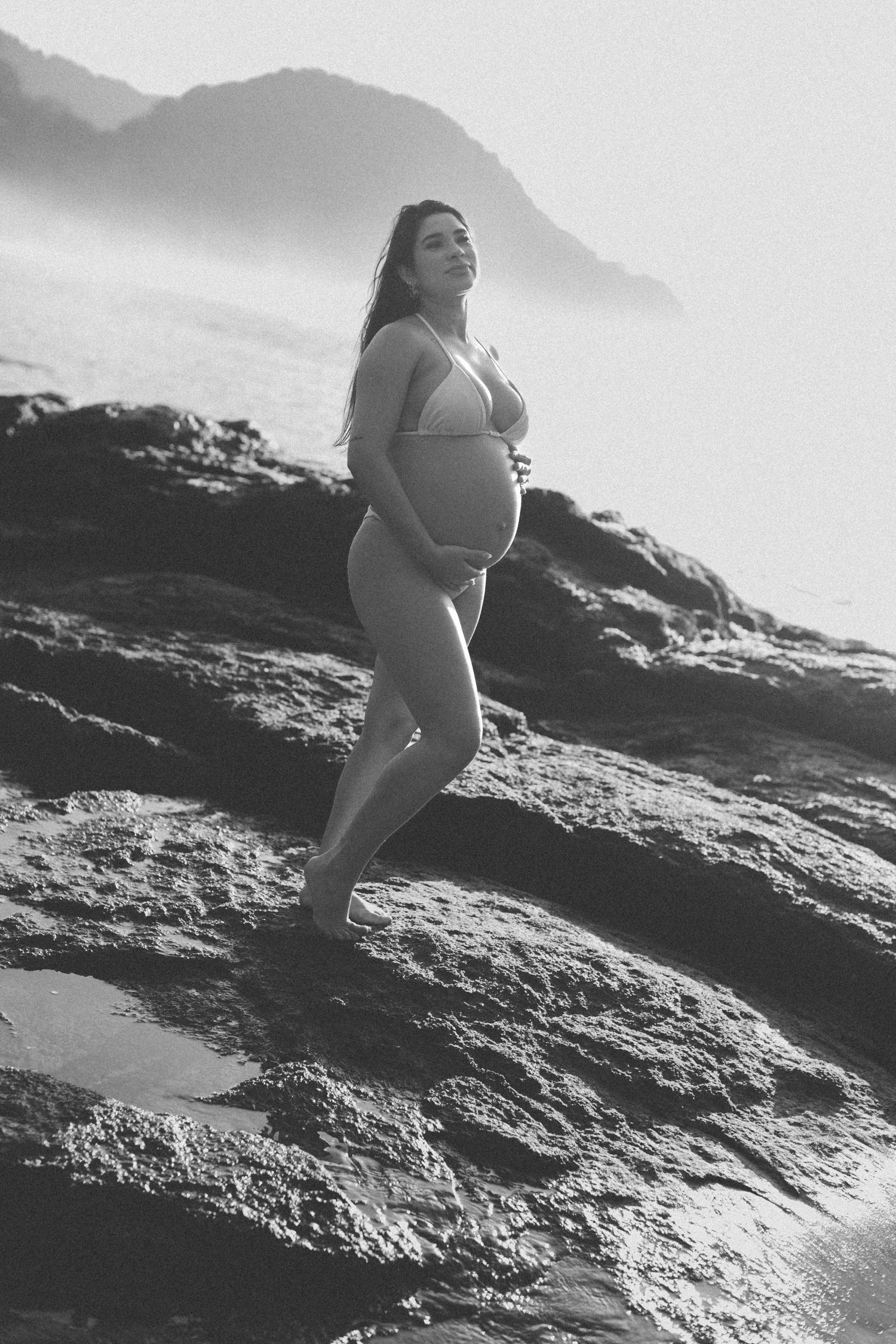 Black and white maternity photo on coastal rocks with mountains in background.