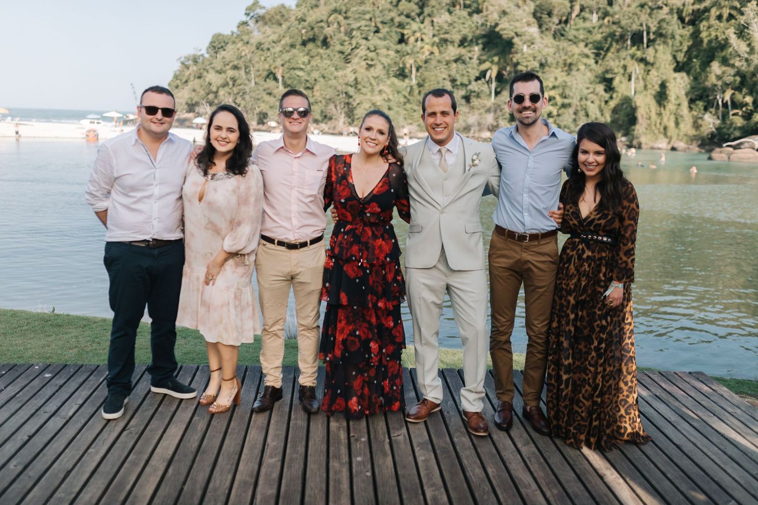 Wedding party in casual attire poses together on wooden deck by the water.