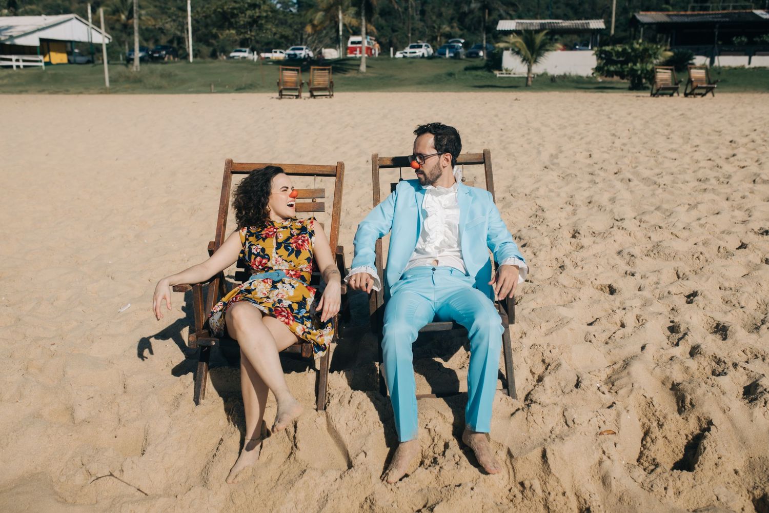 A couple relaxes in wooden beach chairs on sandy shoreline.