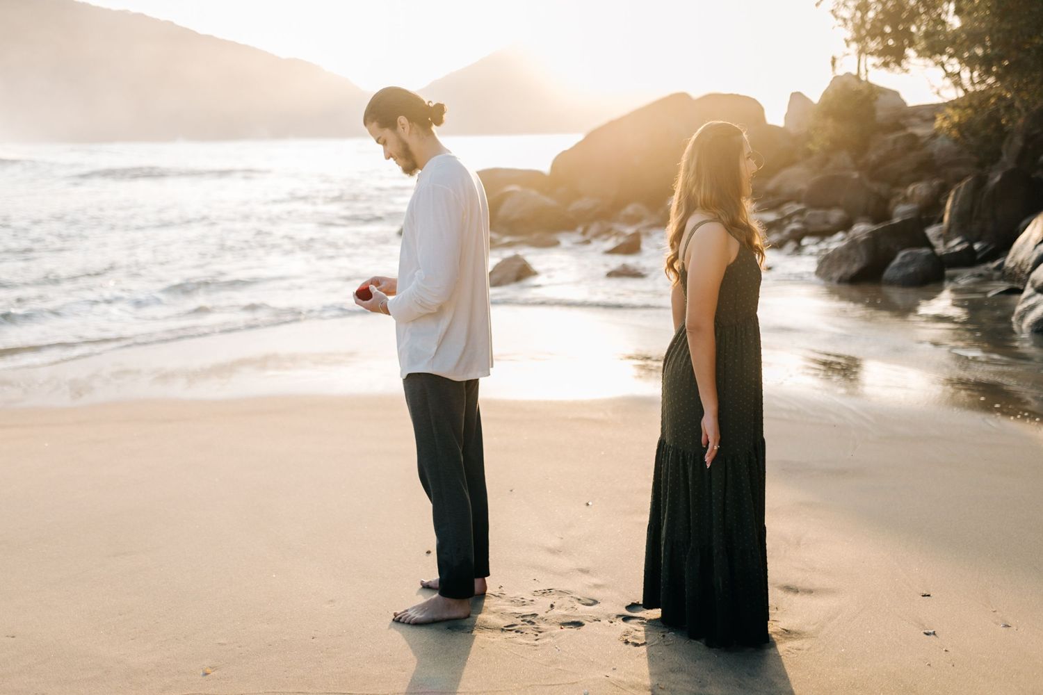 Couple standing apart looking down at beach sand during sunset with waves and rocks in background.