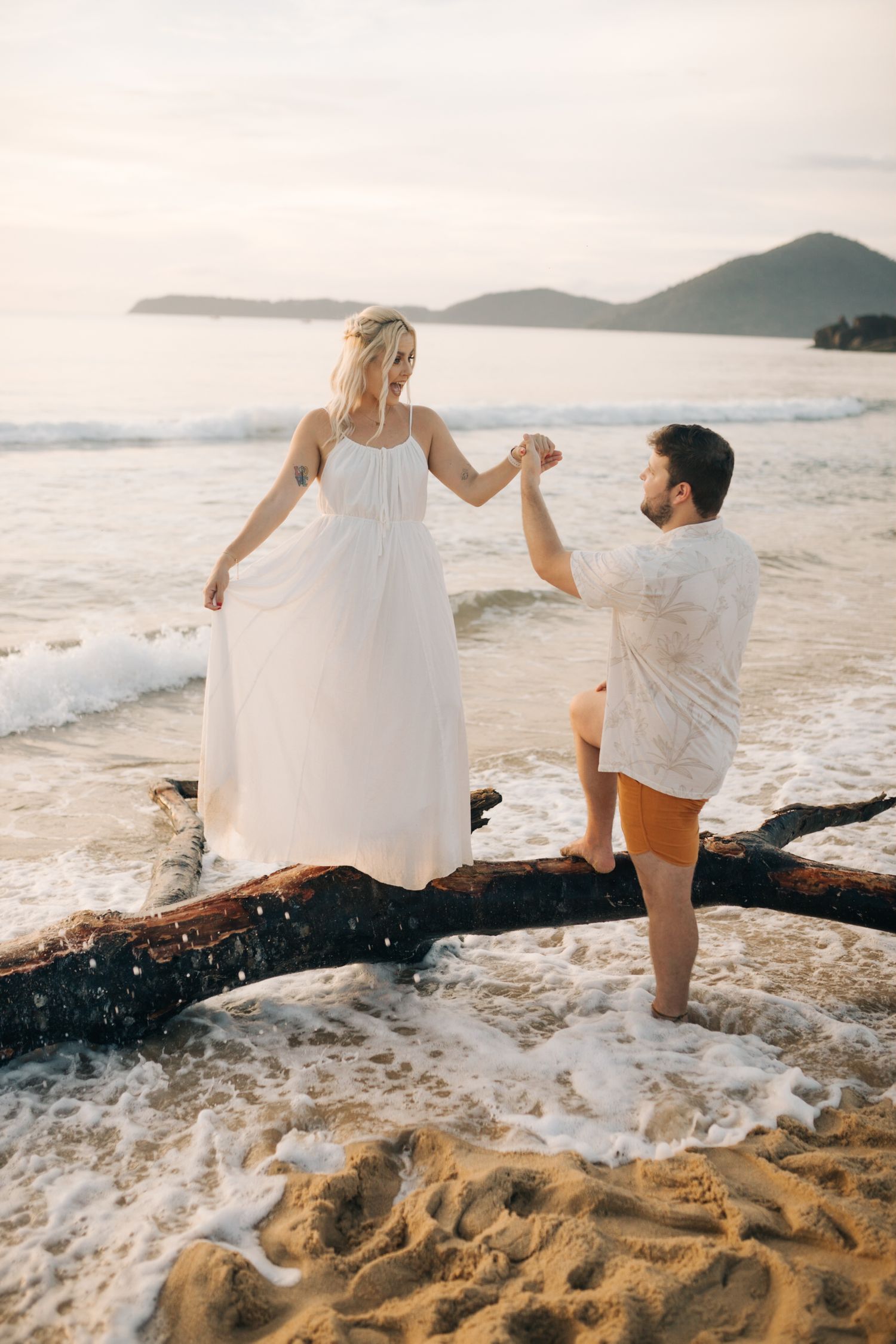 Two people balance on driftwood at the beach as waves crash around them.