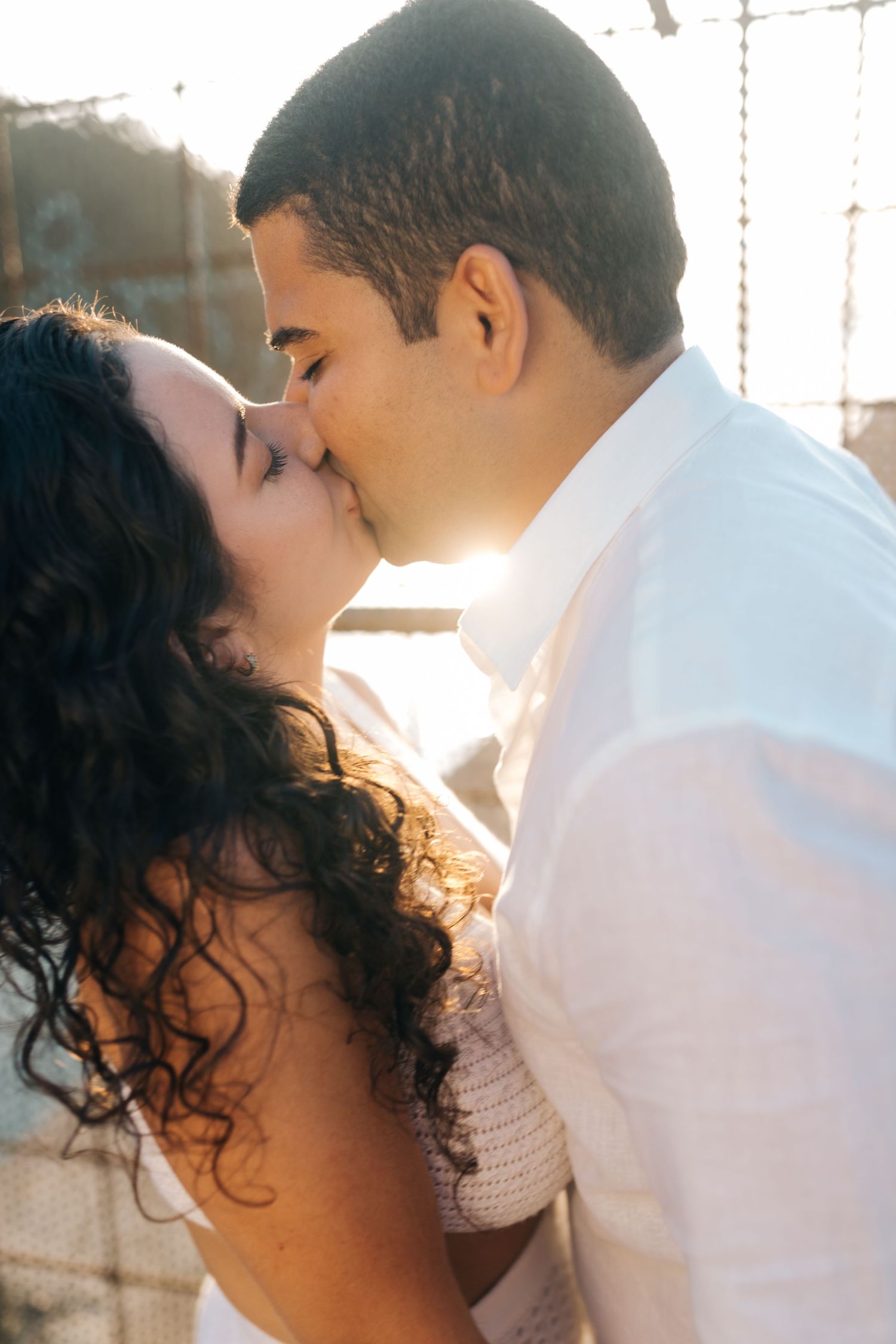 A romantic couple shares a tender kiss in warm sunlit setting wearing white.
