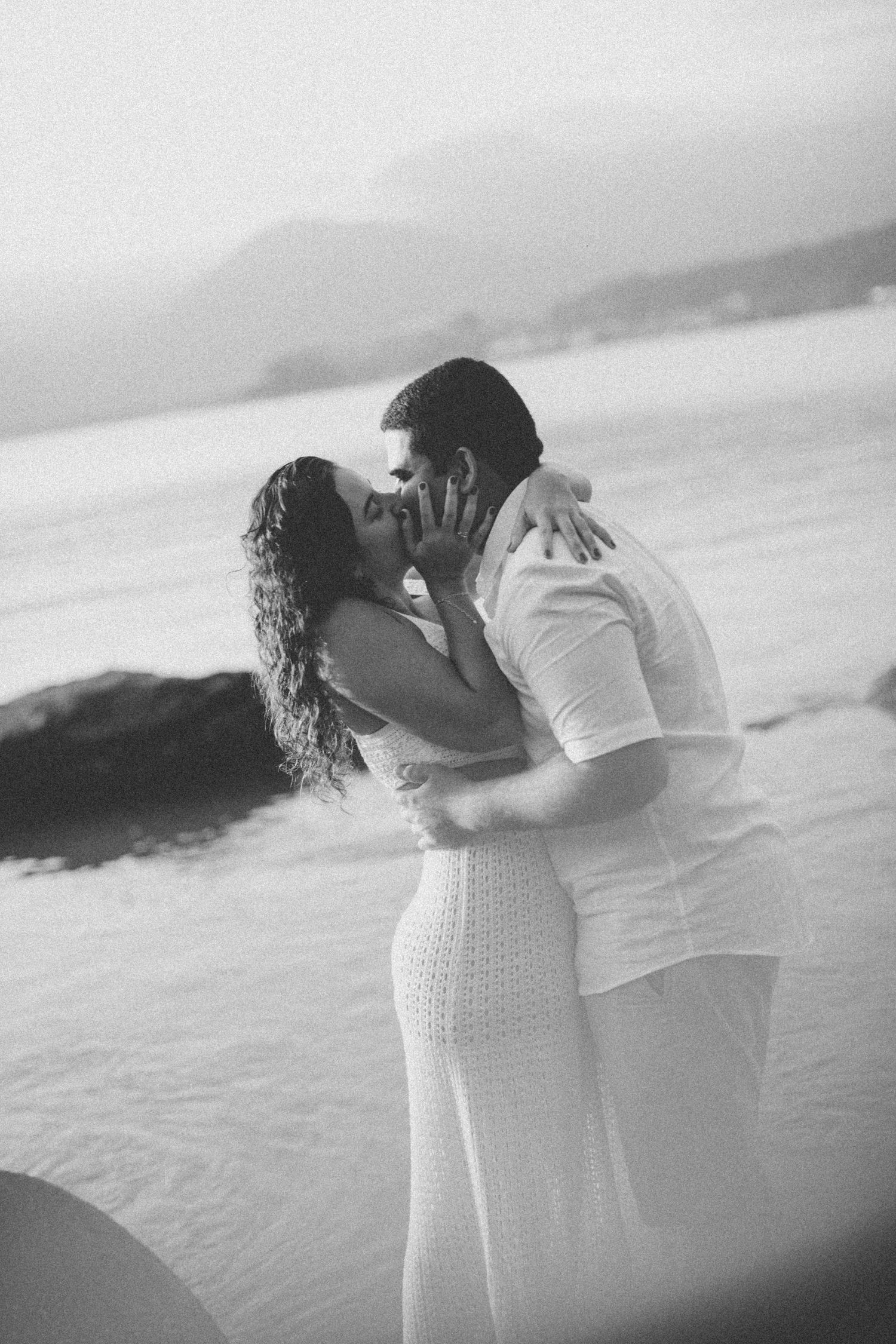 Black and white photo of couple embracing on beach with mountains in background.
