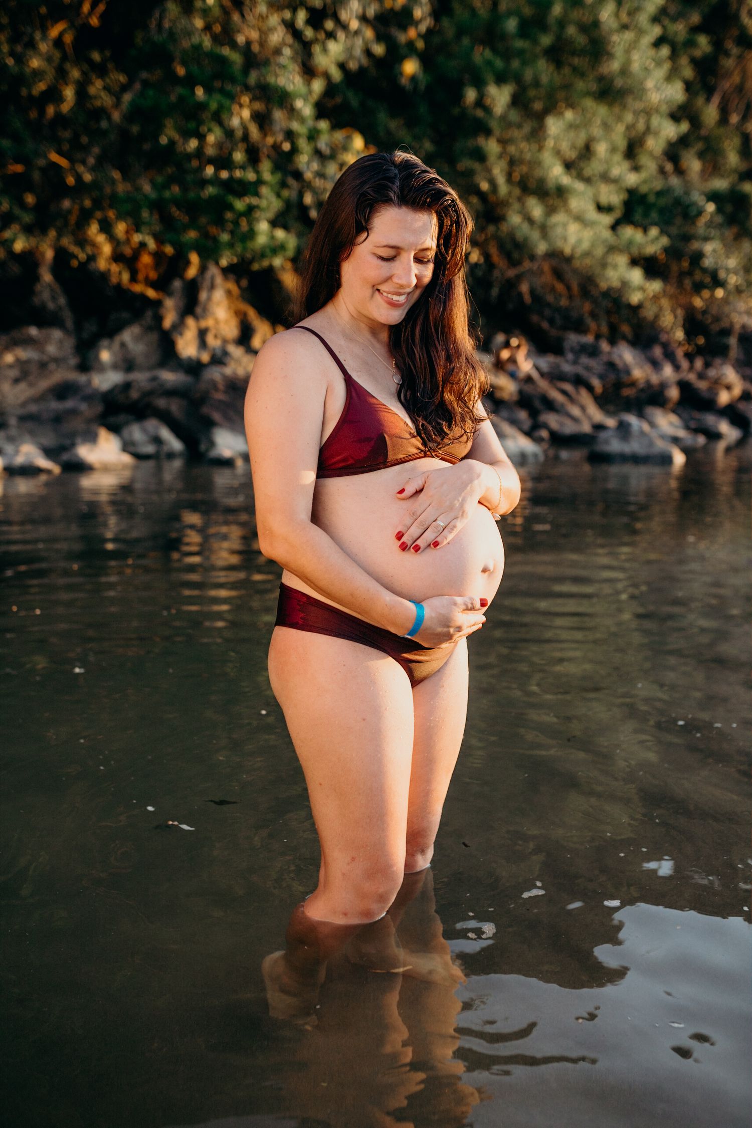 Maternity photos captured at sunset in calm waters near rocky shoreline.