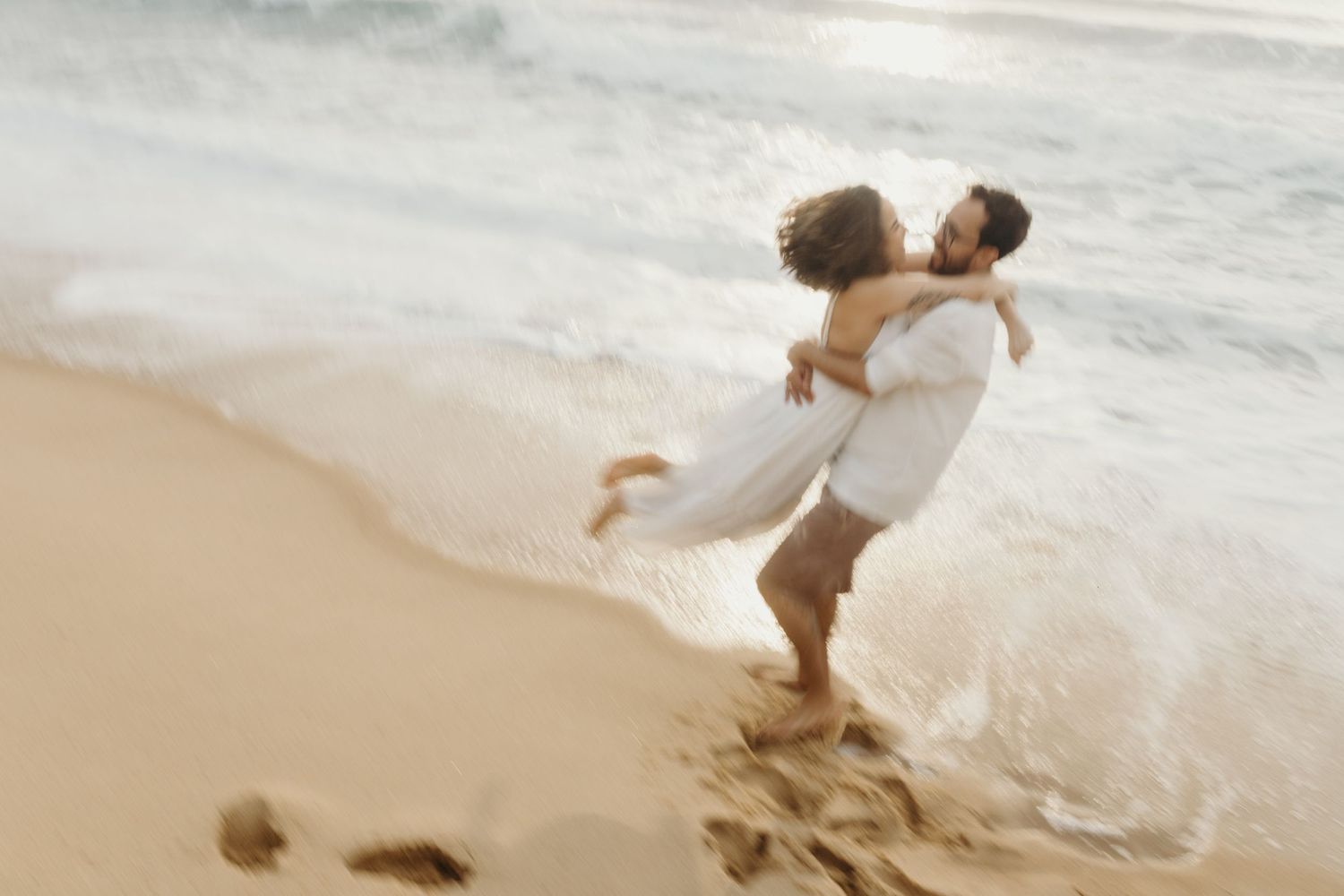 A couple spins joyfully on the beach with waves lapping at their feet.