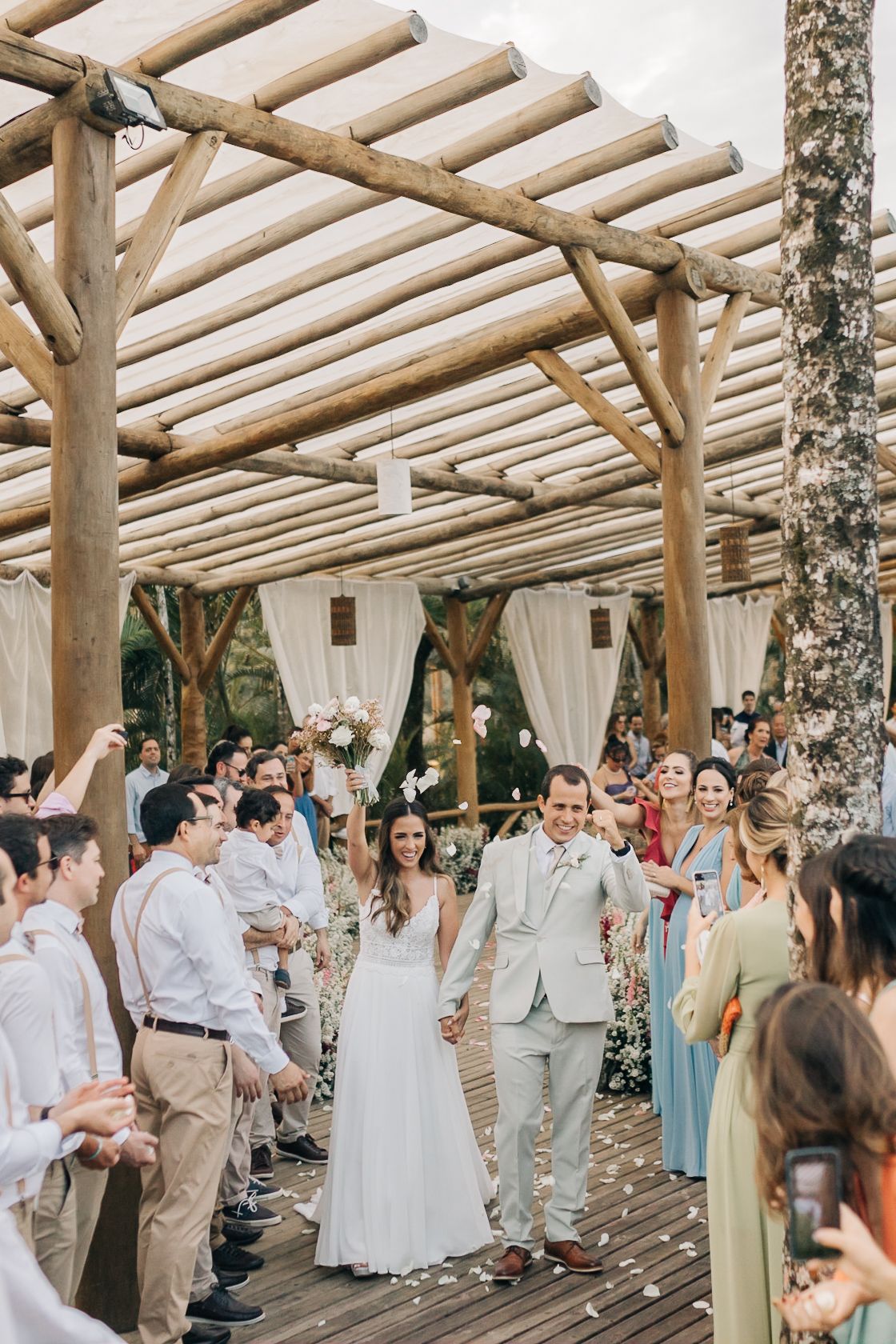 Wedding guests gather under a rustic wooden pergola decorated with white drapes and string lights.