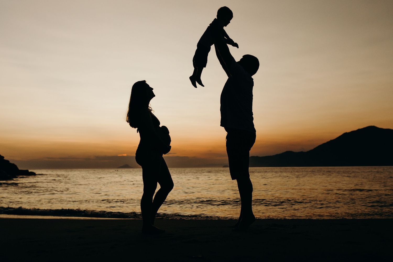 Silhouetted figures share a special family moment together at sunset on a beach with mountains in the background.
