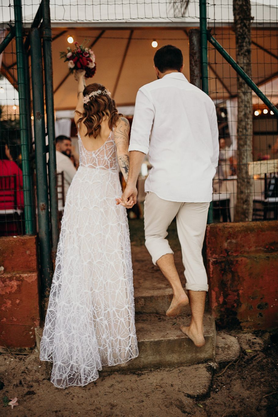 A couple in white outfits walks together during a wedding celebration with decorative lights in the background.