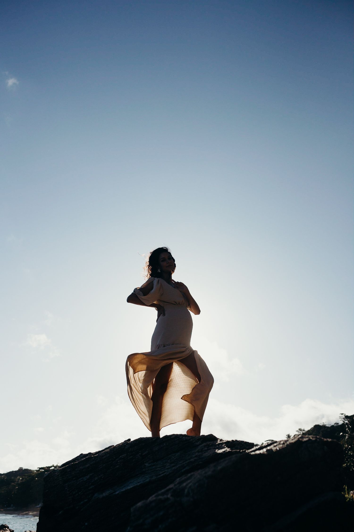 Person in billowing dress stands on rocky outcrop with wind-swept fabric against clear sky.