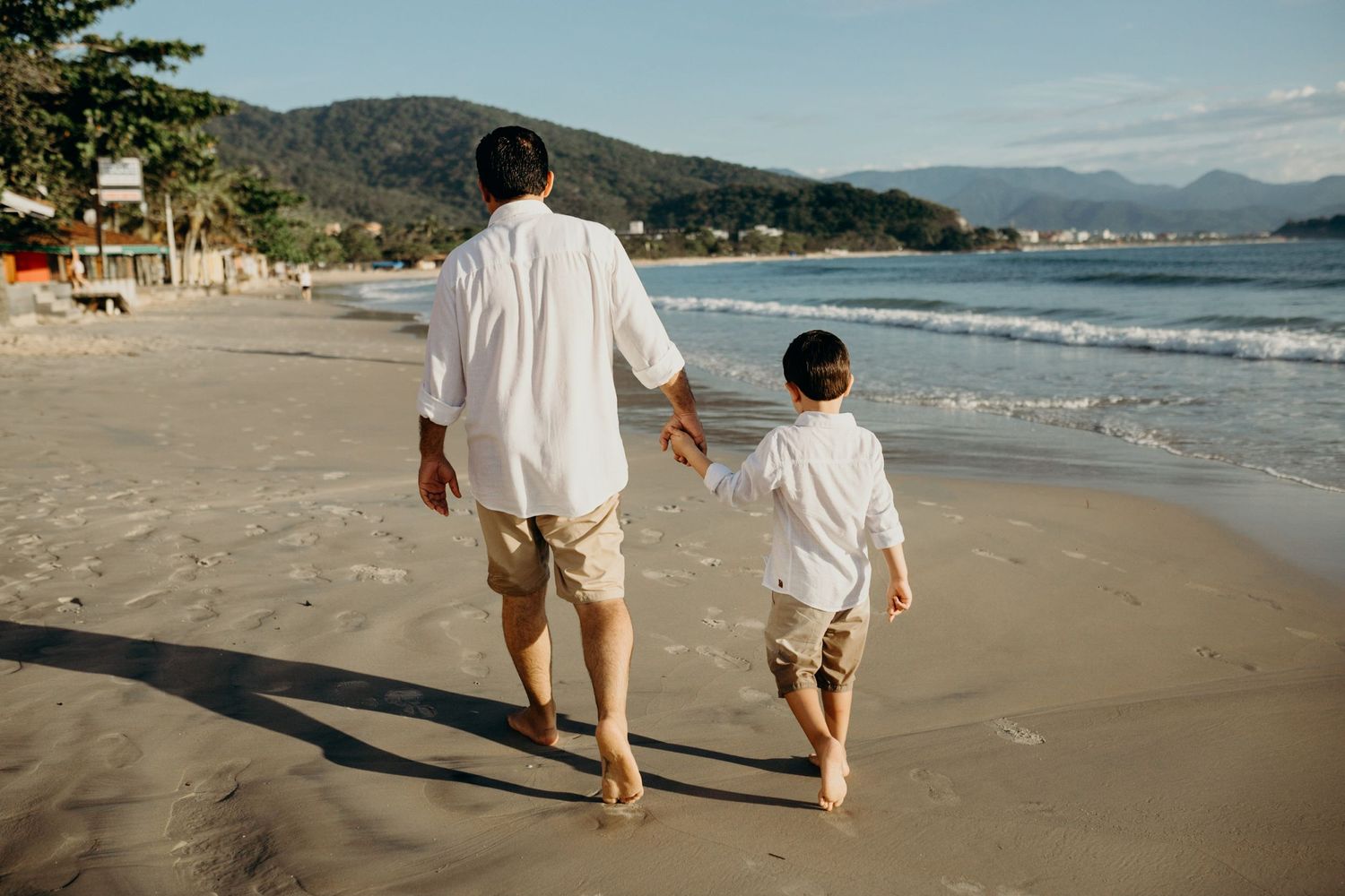Two figures walking together barefoot on a scenic beach with mountains in the background.