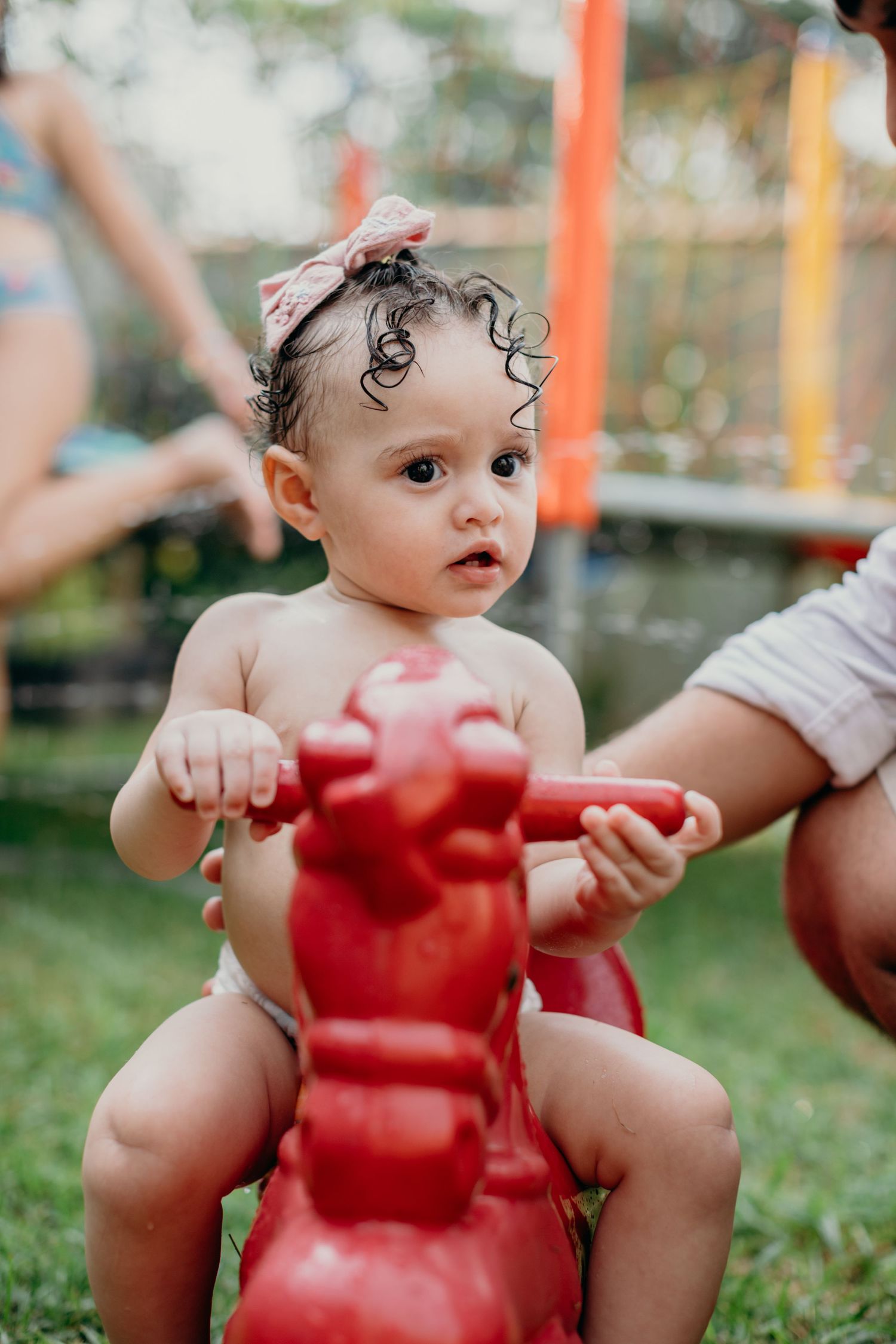 A toddler in a headband sits on a red rocking horse toy in an outdoor setting.