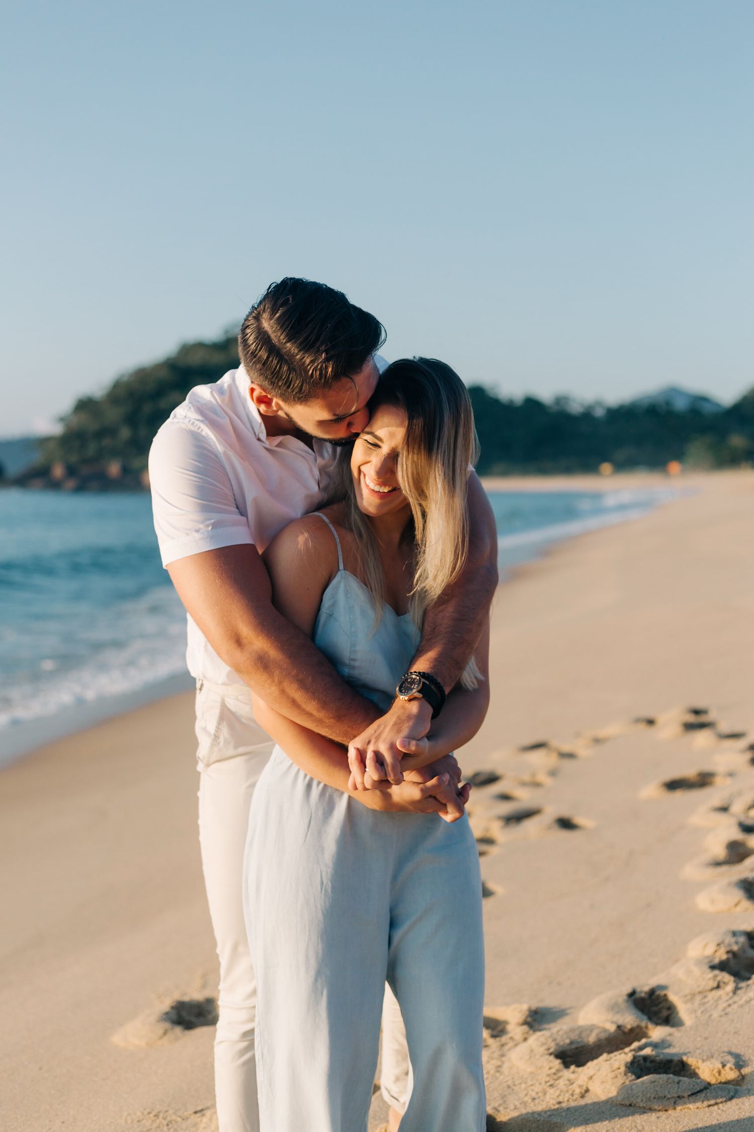 Loving couple in white outfits embracing on sunlit beach with coastal scenery.