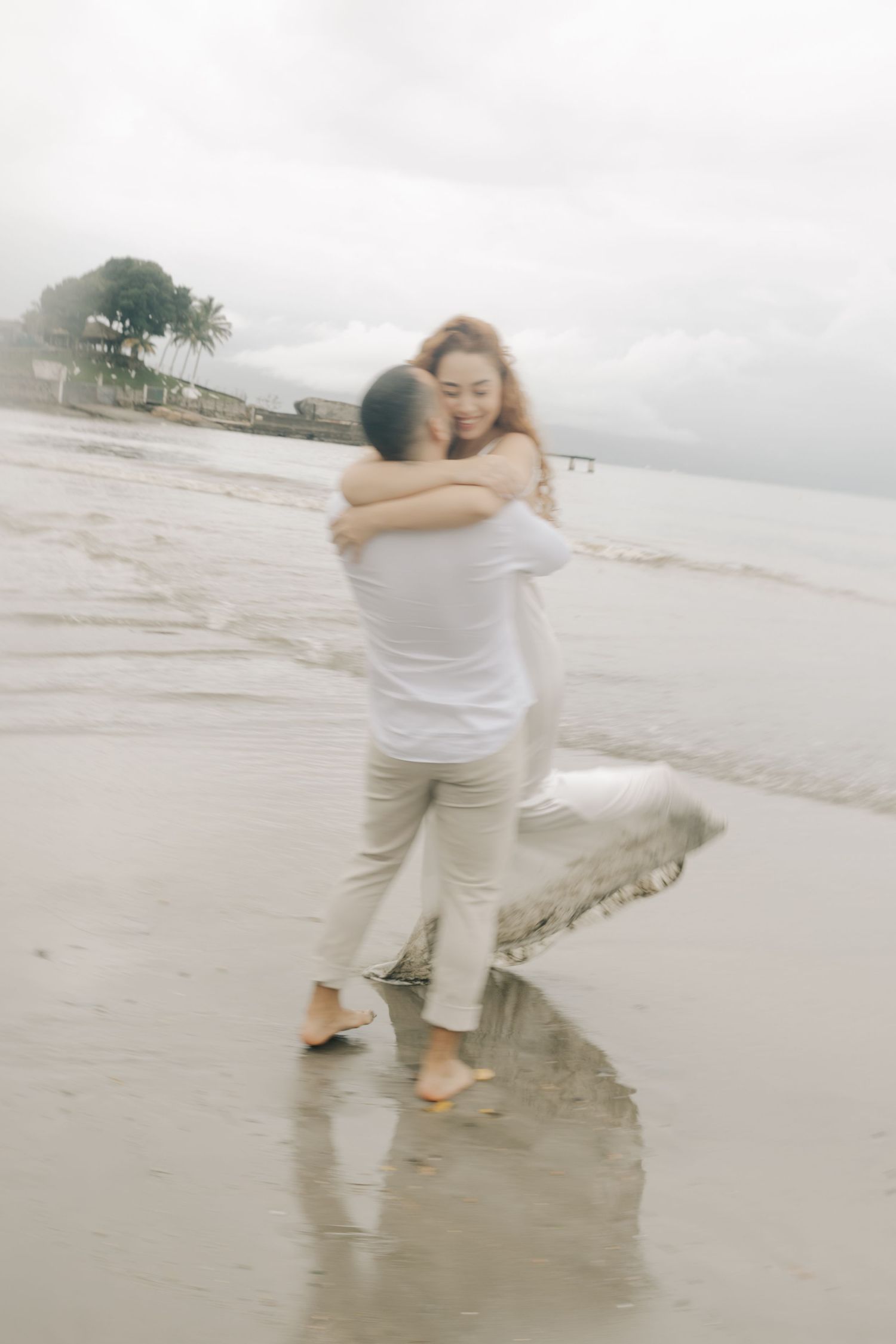 A couple in white clothing spins and dances together on wet beach sand.