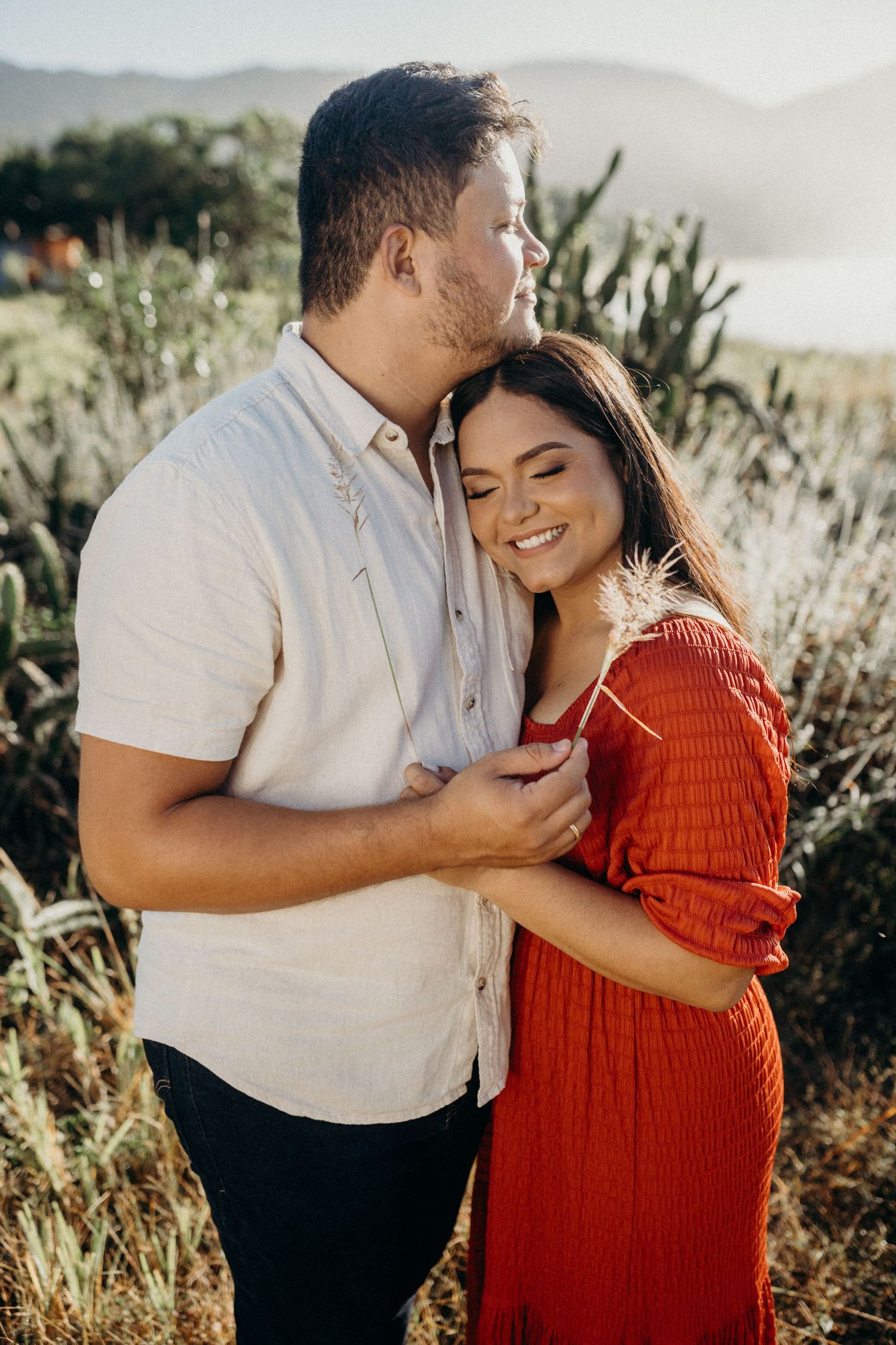 Couple shares tender moment in white and red outfits against mountainous backdrop at sunset.