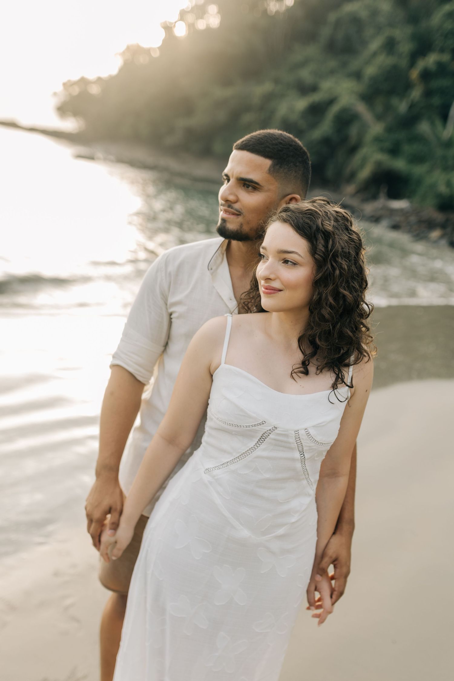 A couple poses intimately at sunset on a beach with water and trees in the background.