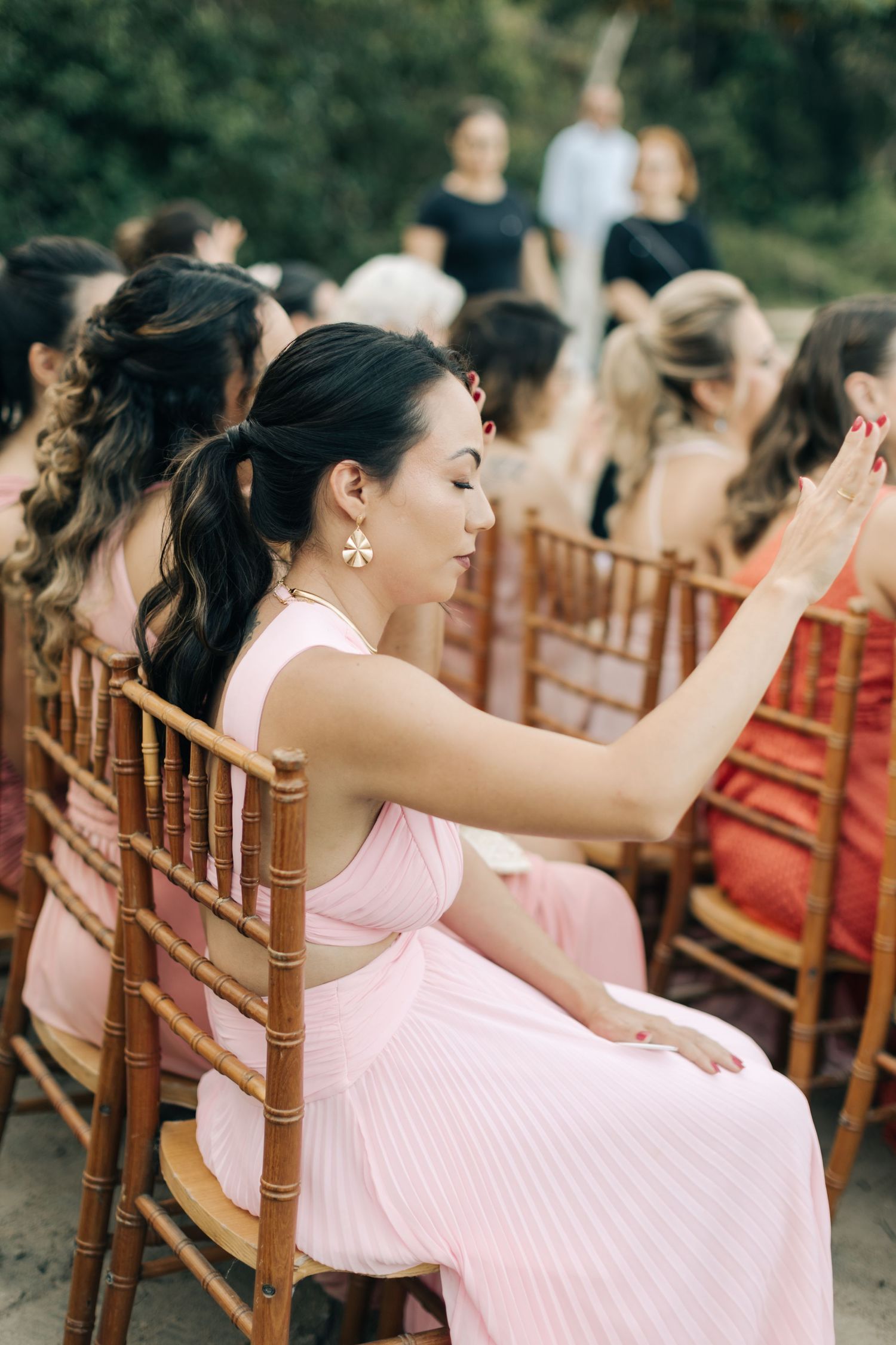 Wedding guests in pink dresses seated in wooden chairs during an outdoor ceremony.