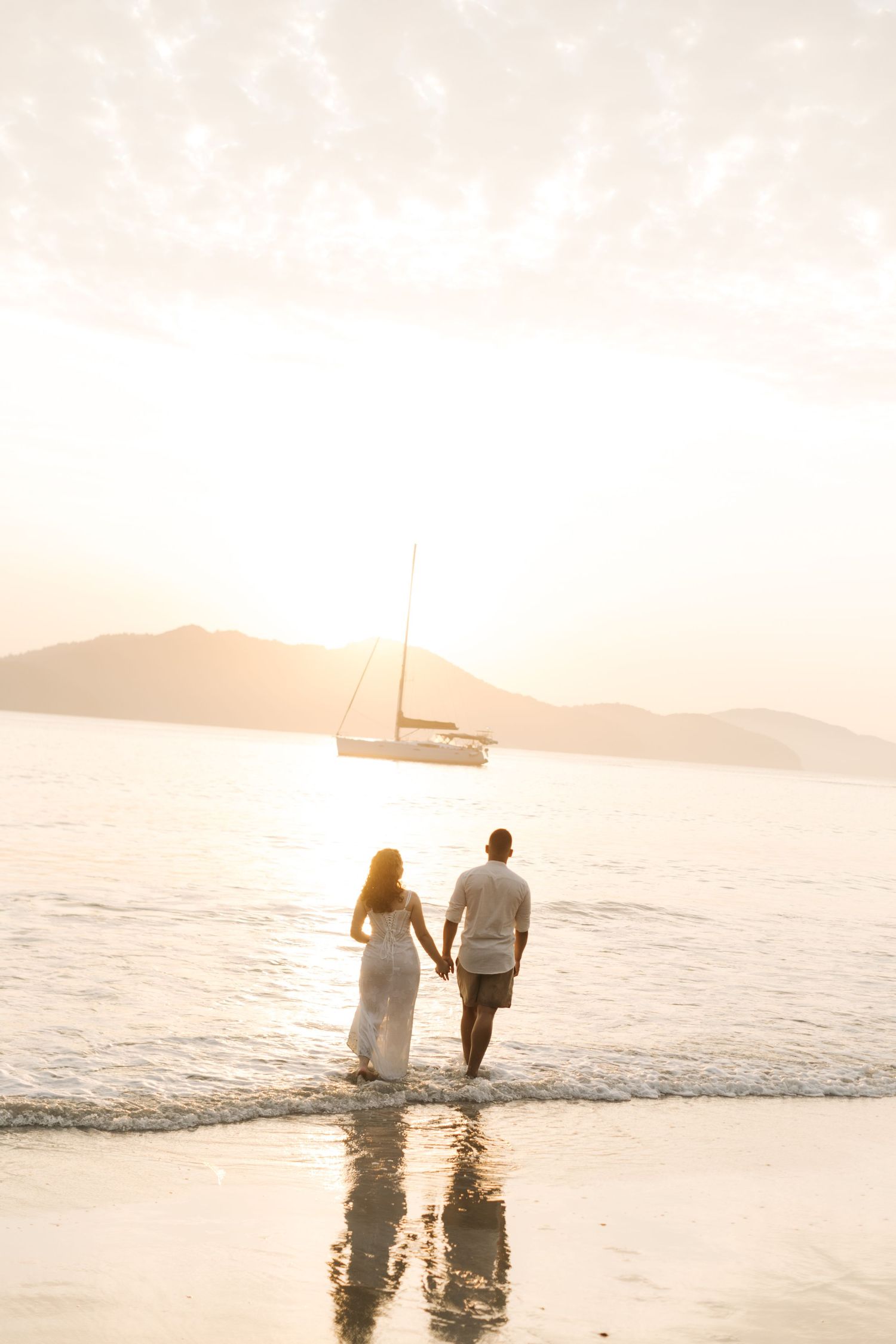 Dreamy beach scene with couple walking through water at sunset and sailboat in distance.