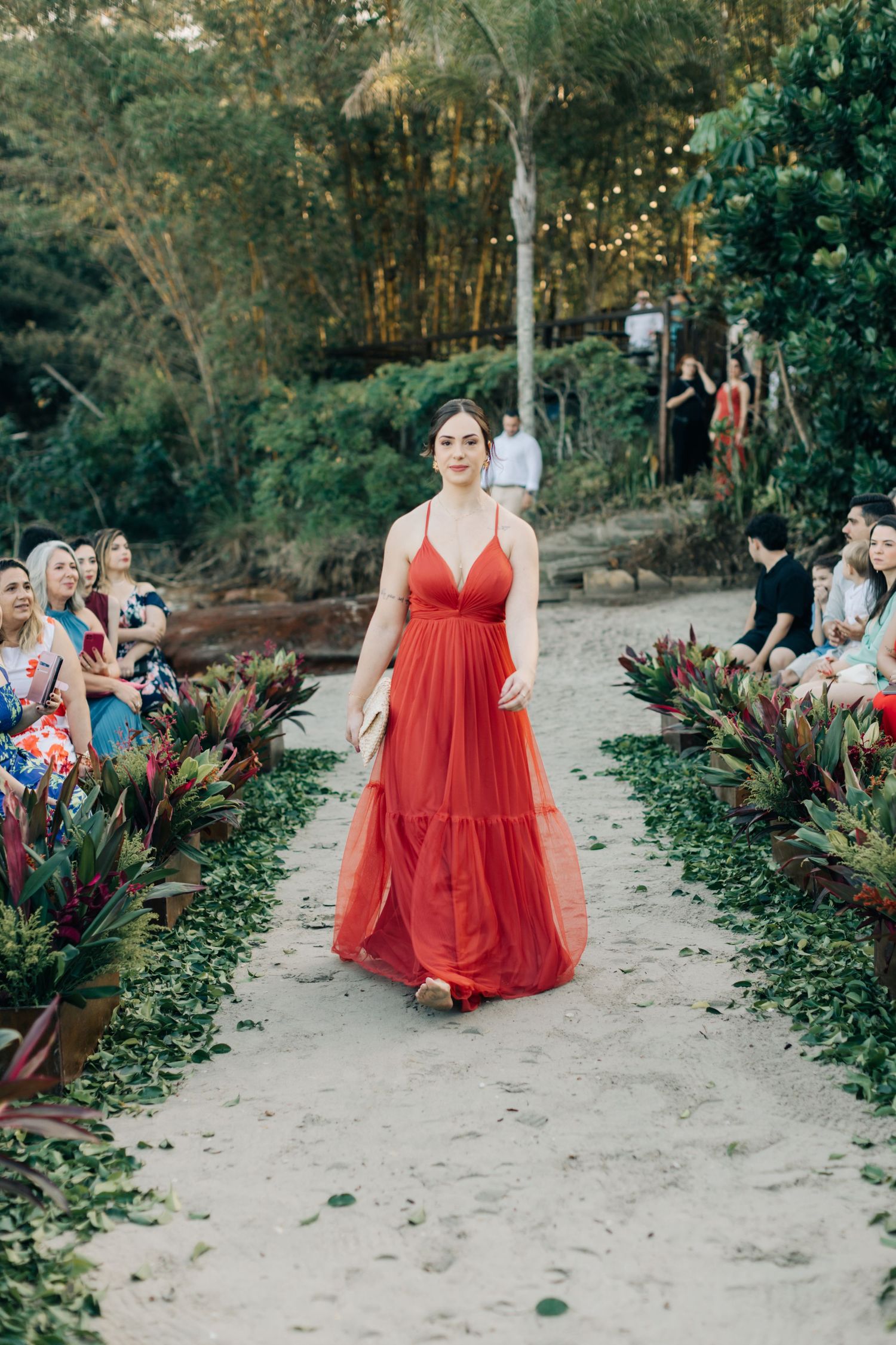 Bridesmaid in flowing red gown walks down garden path at outdoor wedding ceremony.