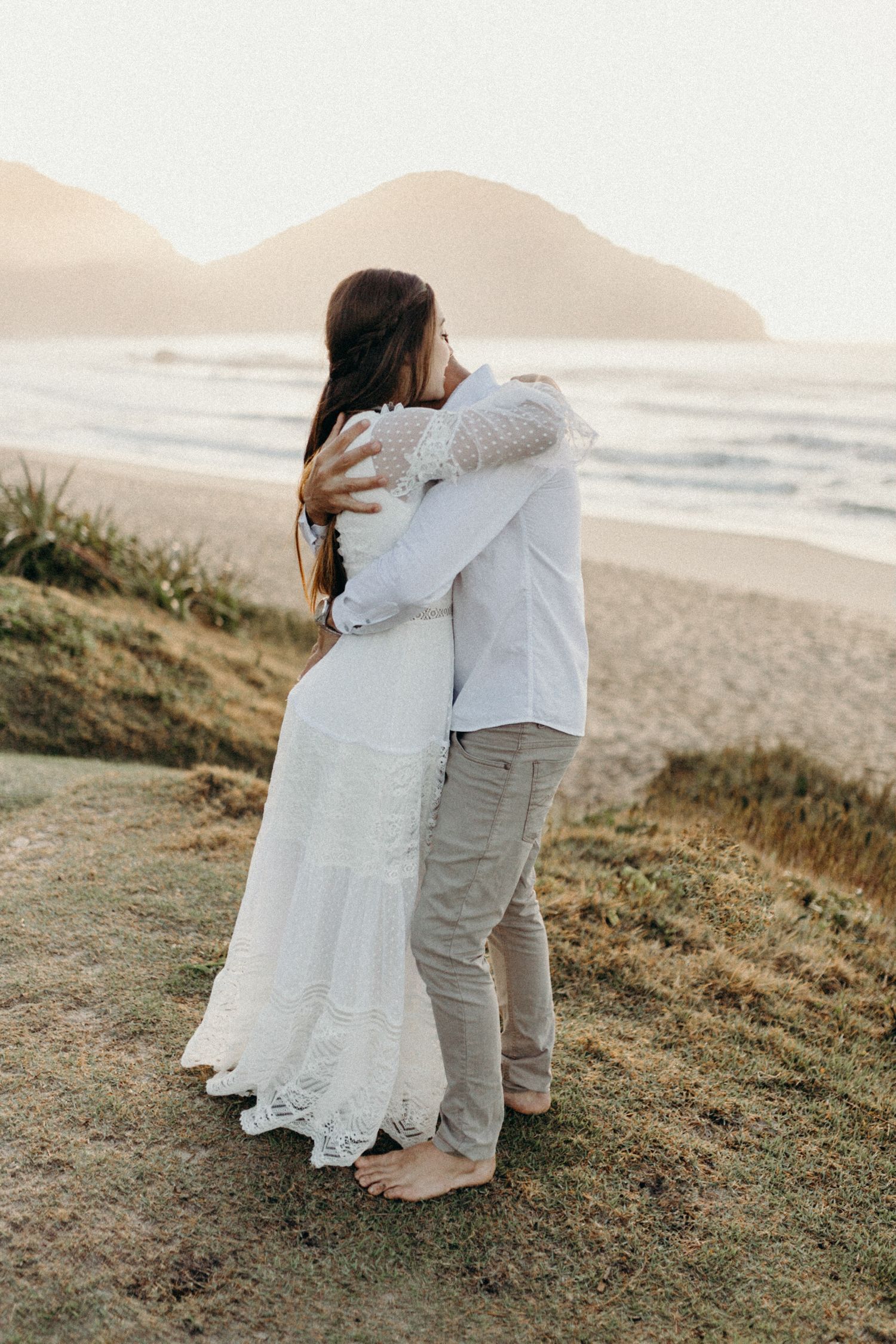 Couple embracing on grassy hillside overlooking beach at sunset.