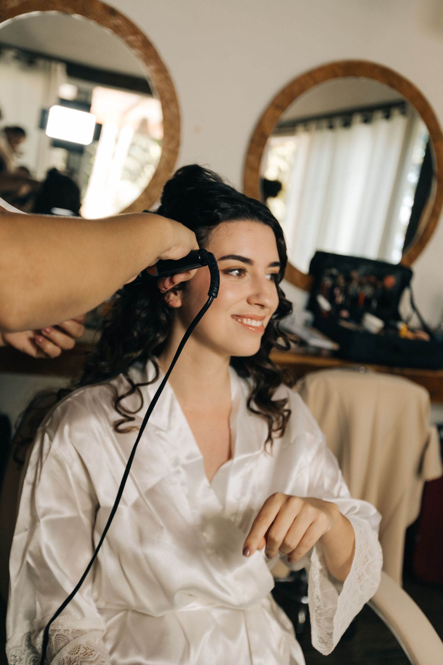 Someone getting their dark curly hair styled in a white robe at a vanity with round mirrors.