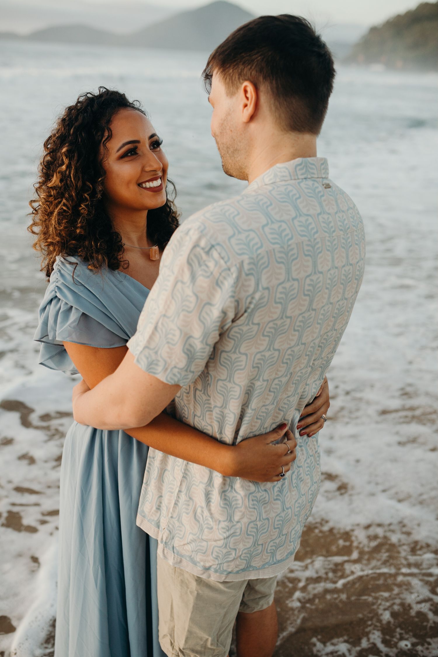 Romantic beach photoshoot with couple embracing by ocean waves during sunset.