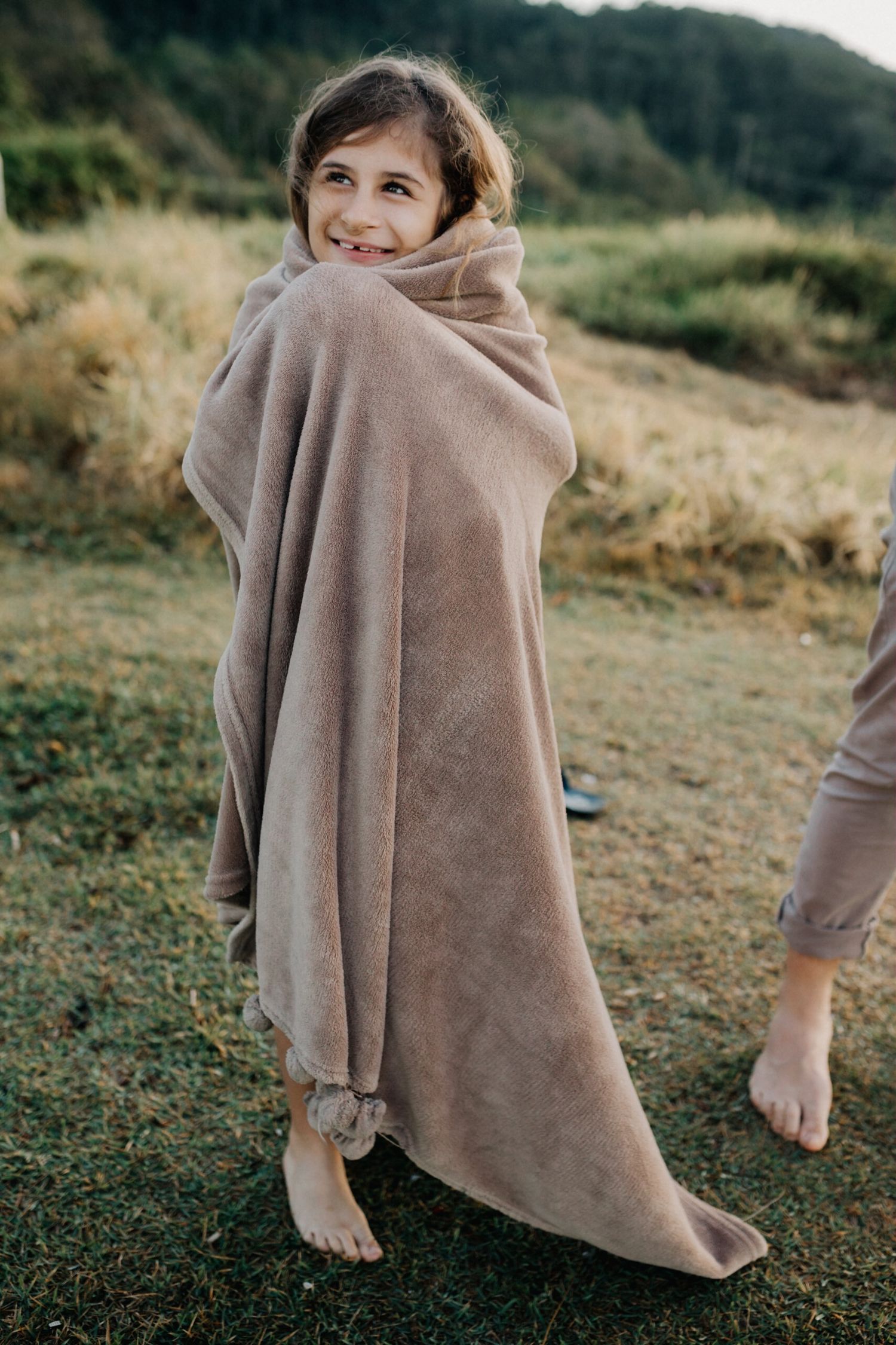 Someone draped in a soft beige blanket posing playfully against a natural outdoor backdrop.